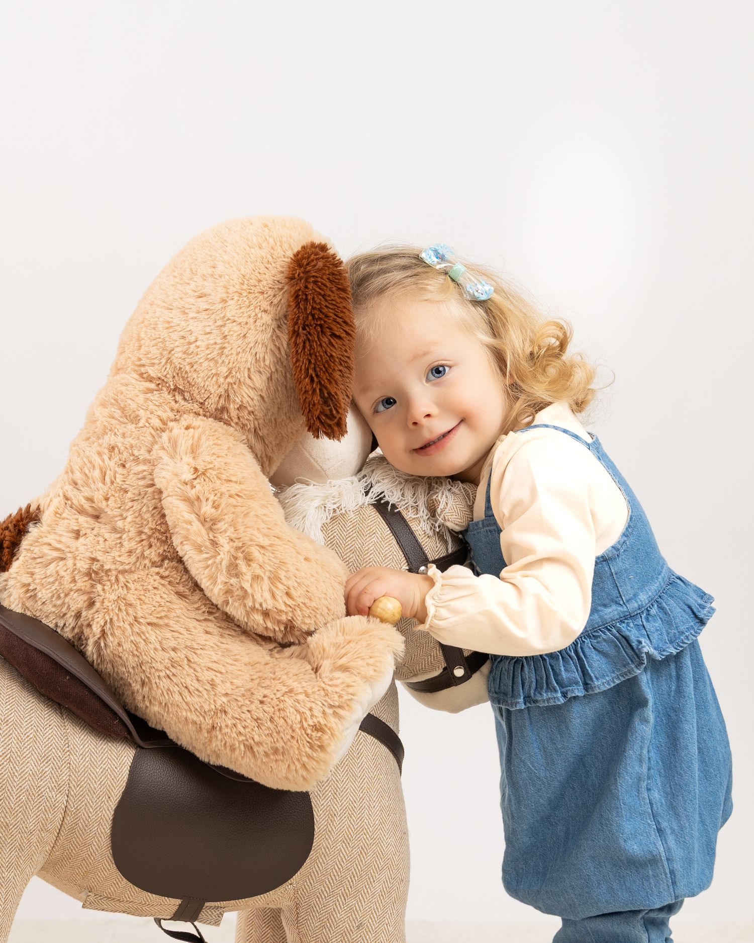 A small child in a denim dress hugs a large beige teddy bear in a series of sweet candid moments.