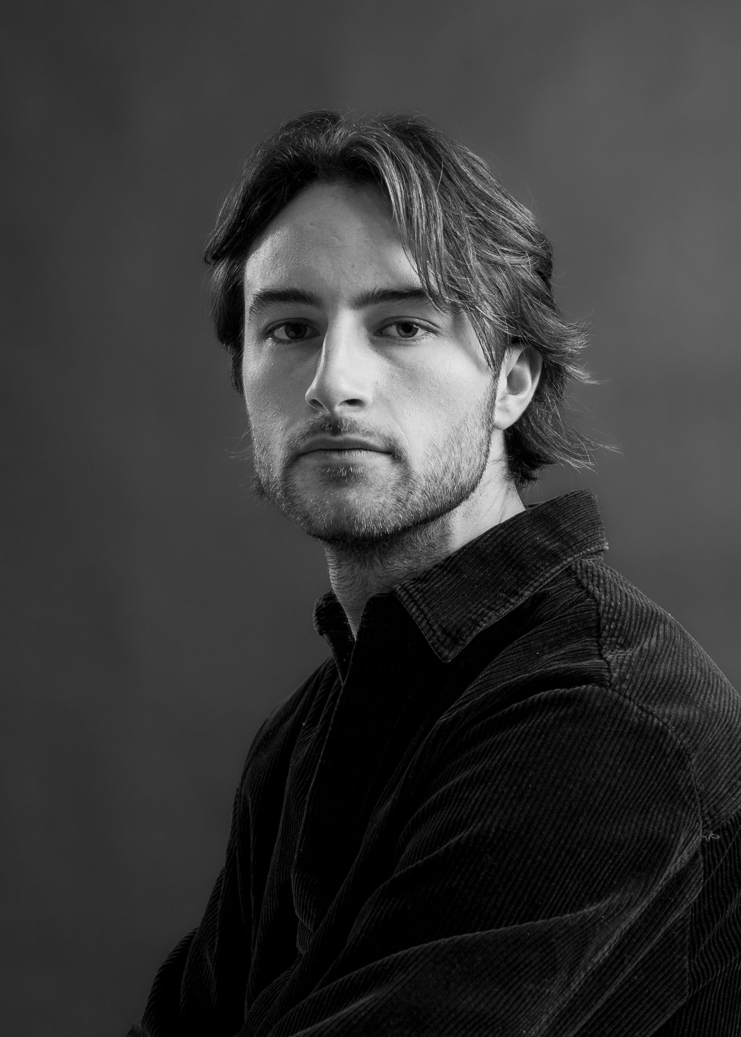 Black and white portrait photograph of a person wearing a dark corduroy jacket against a shadowy background. Creative headshot