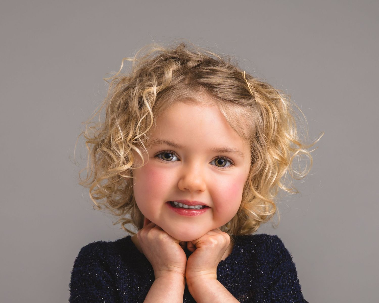 Adorable toddler girl with curly blonde hair smiling with hands under chin against gray background.