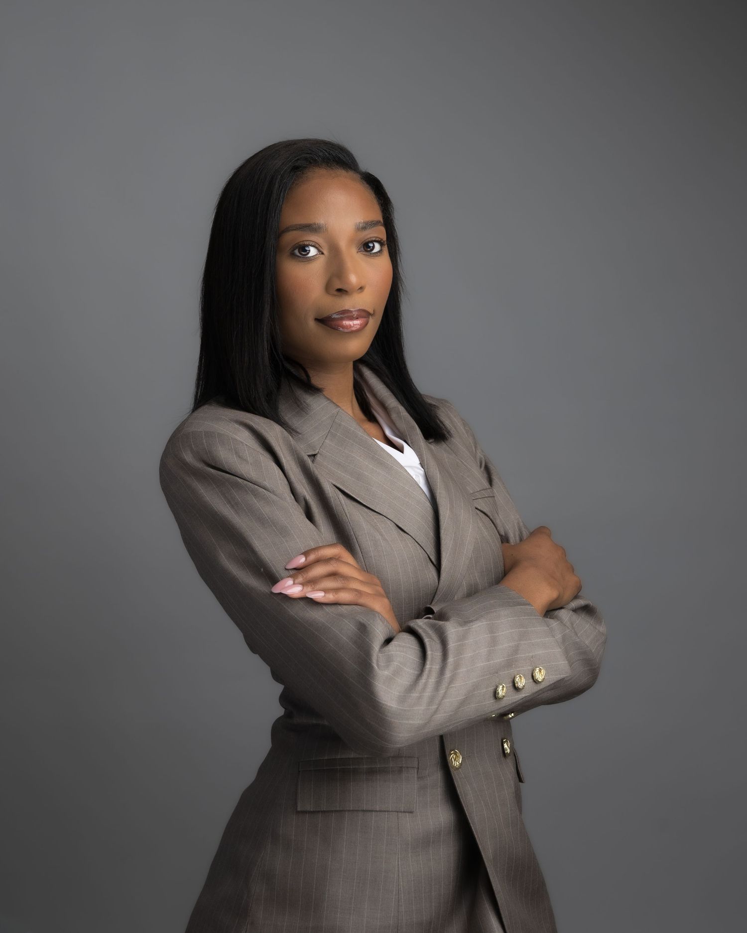 Professional Black woman in gray blazer with arms crossed, confident business portrait on gray background.