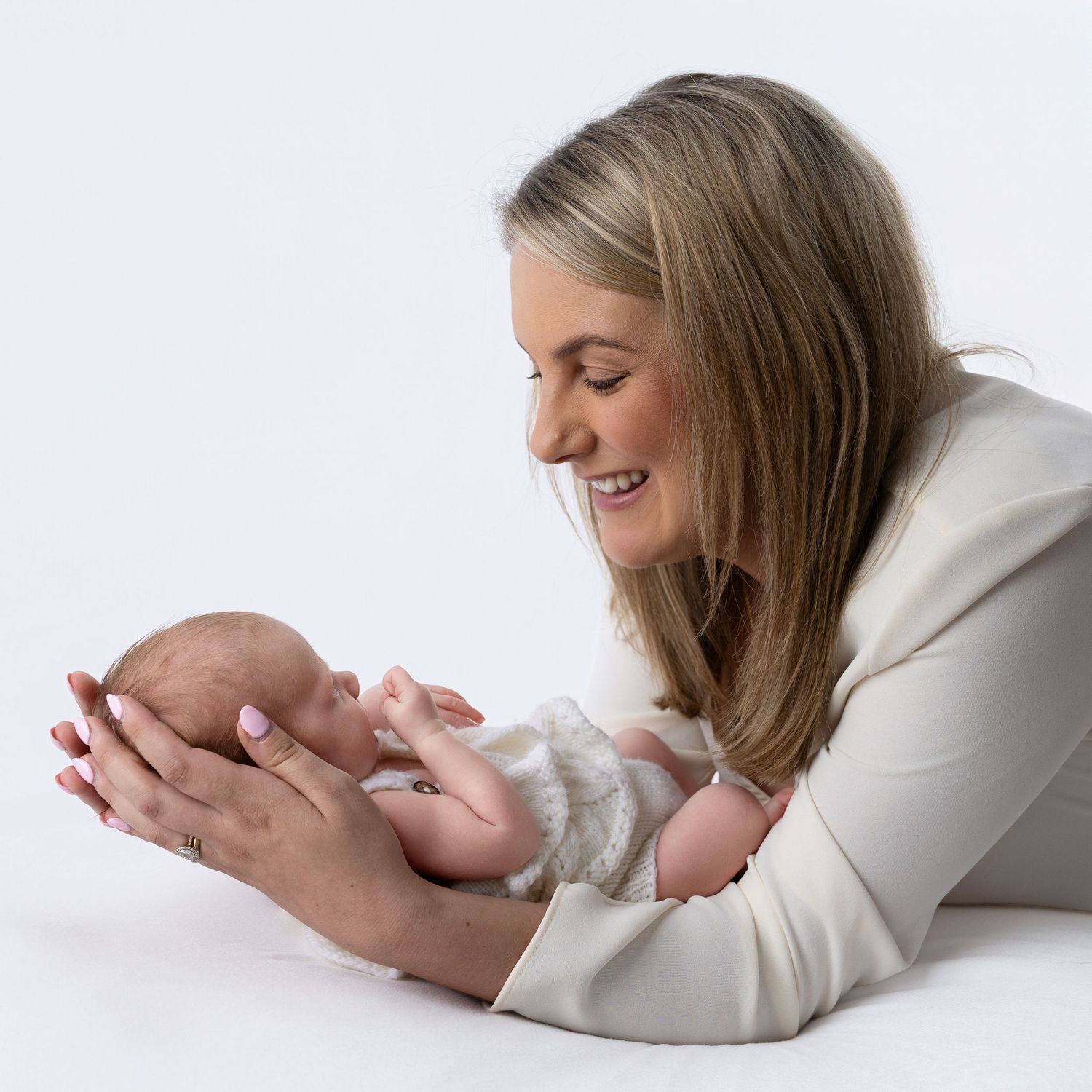 A smiling mother in a cream outfit tenderly holds her sleeping newborn baby against a white background.