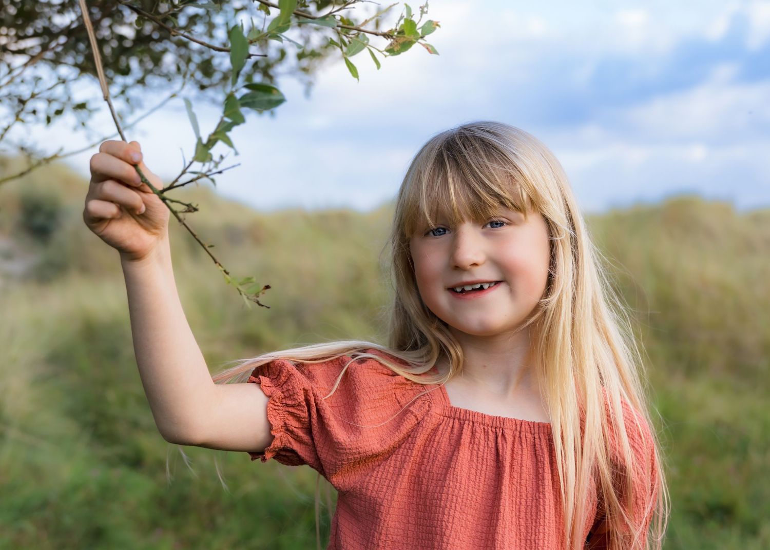 Child in coral dress holding up green plant branch against blue sky.