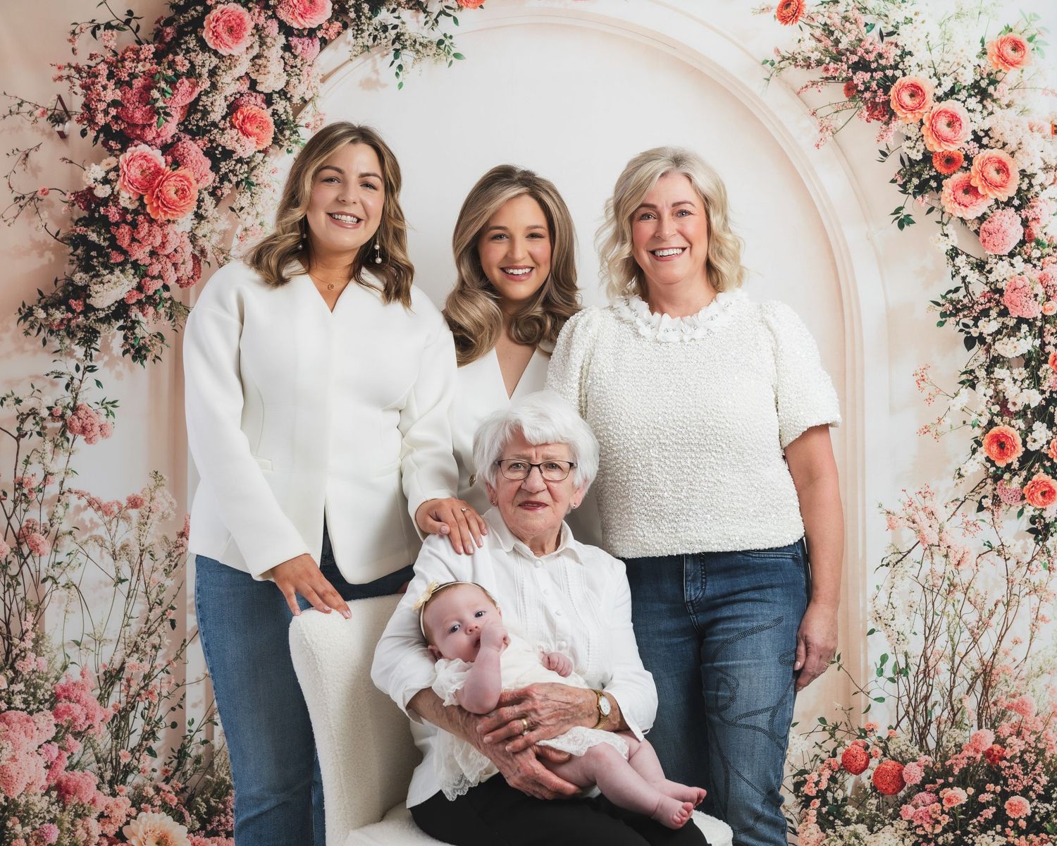 Four generations of family members pose together against a floral backdrop with pink roses and greenery.