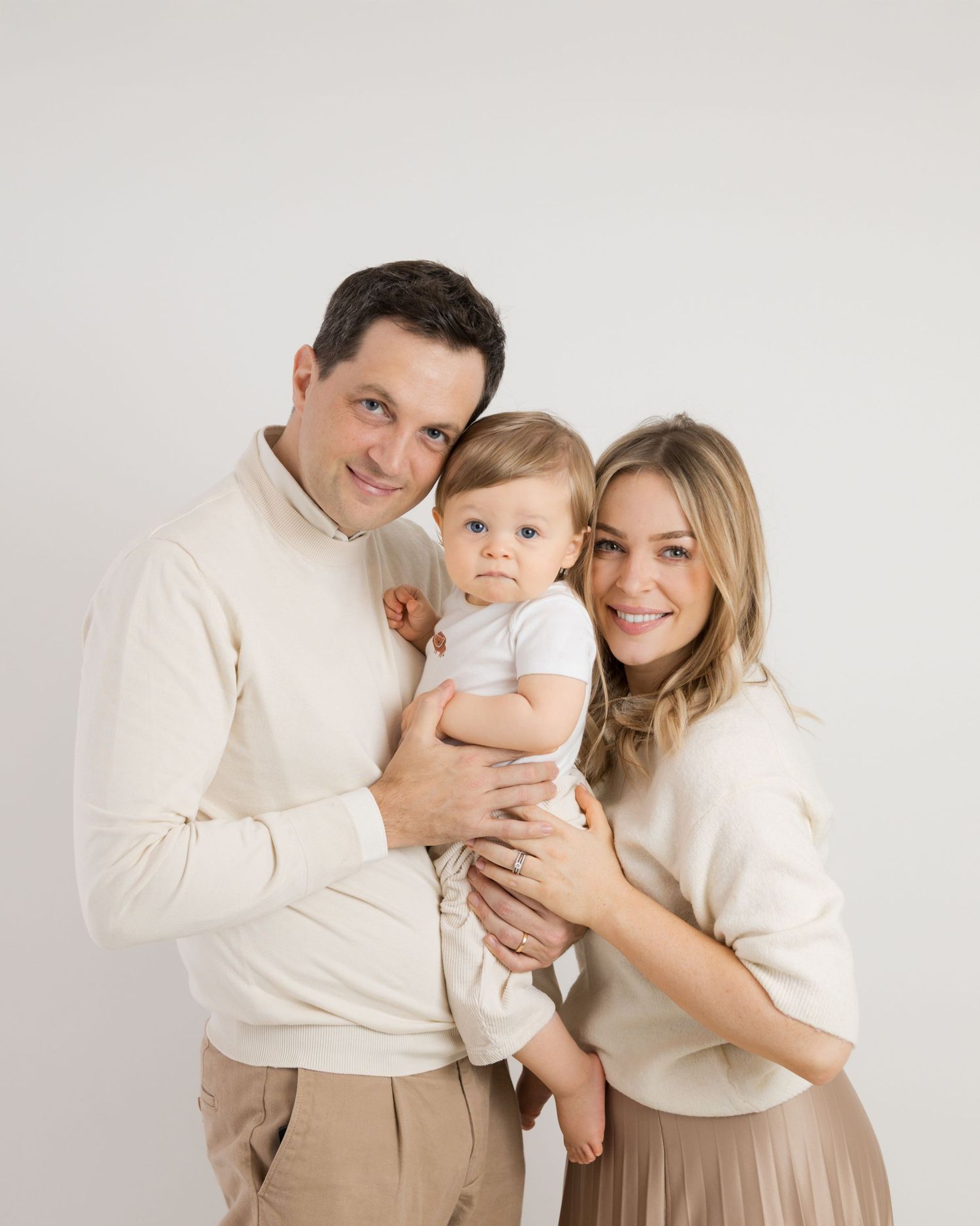 Family wearing cream colored sweaters poses together against white background for studio portraits.