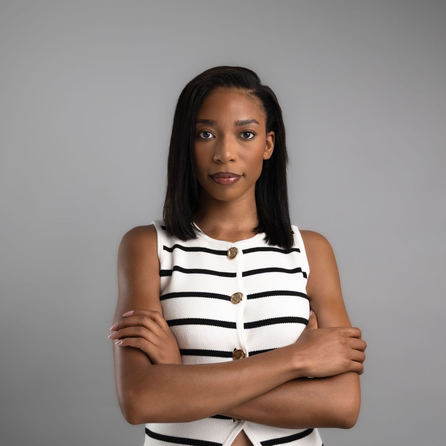 Confident young Black woman in striped sleeveless top, arms crossed, posing against gray background.