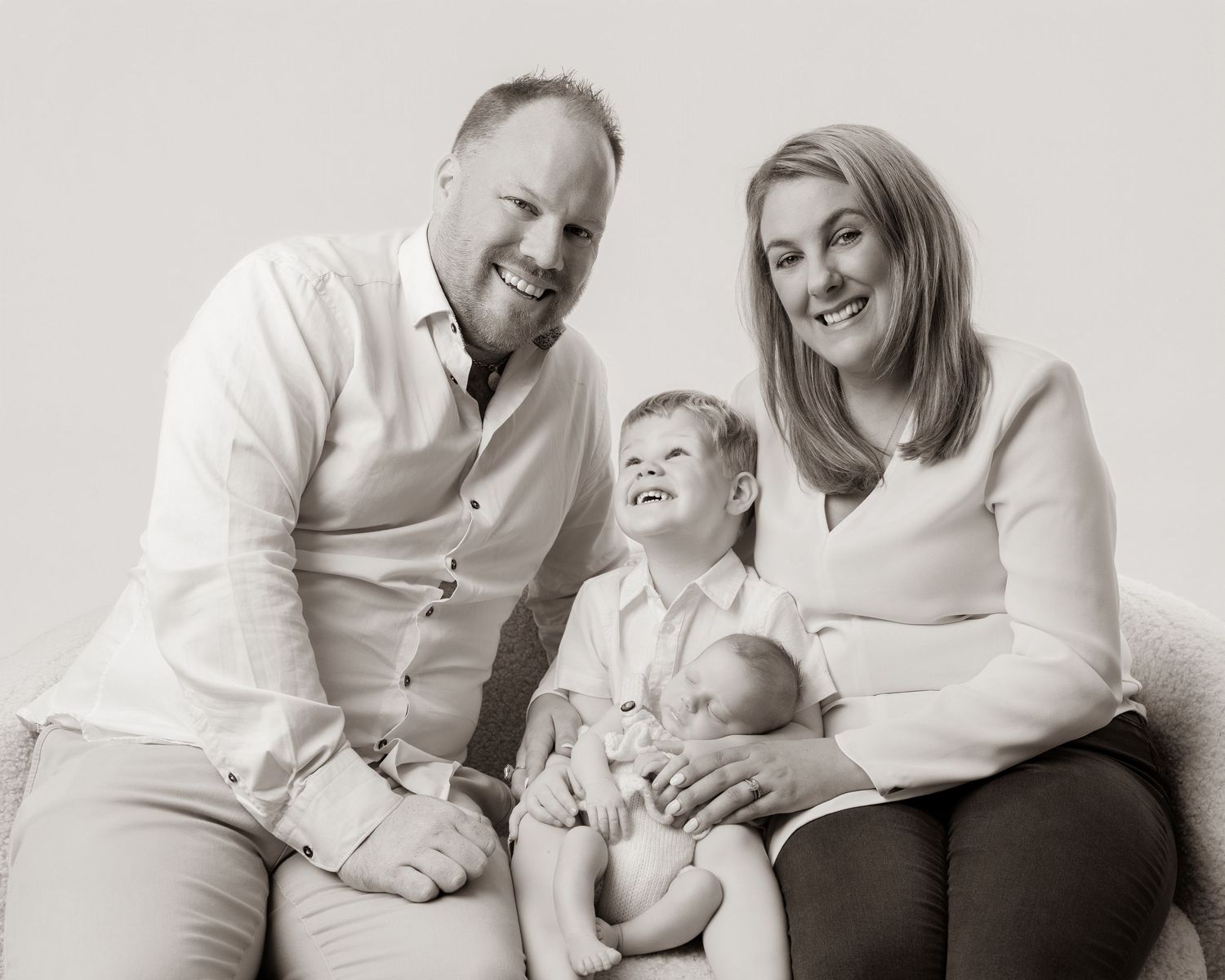 Black and white family portrait of parents with young boy and newborn baby sitting together on a sofa.