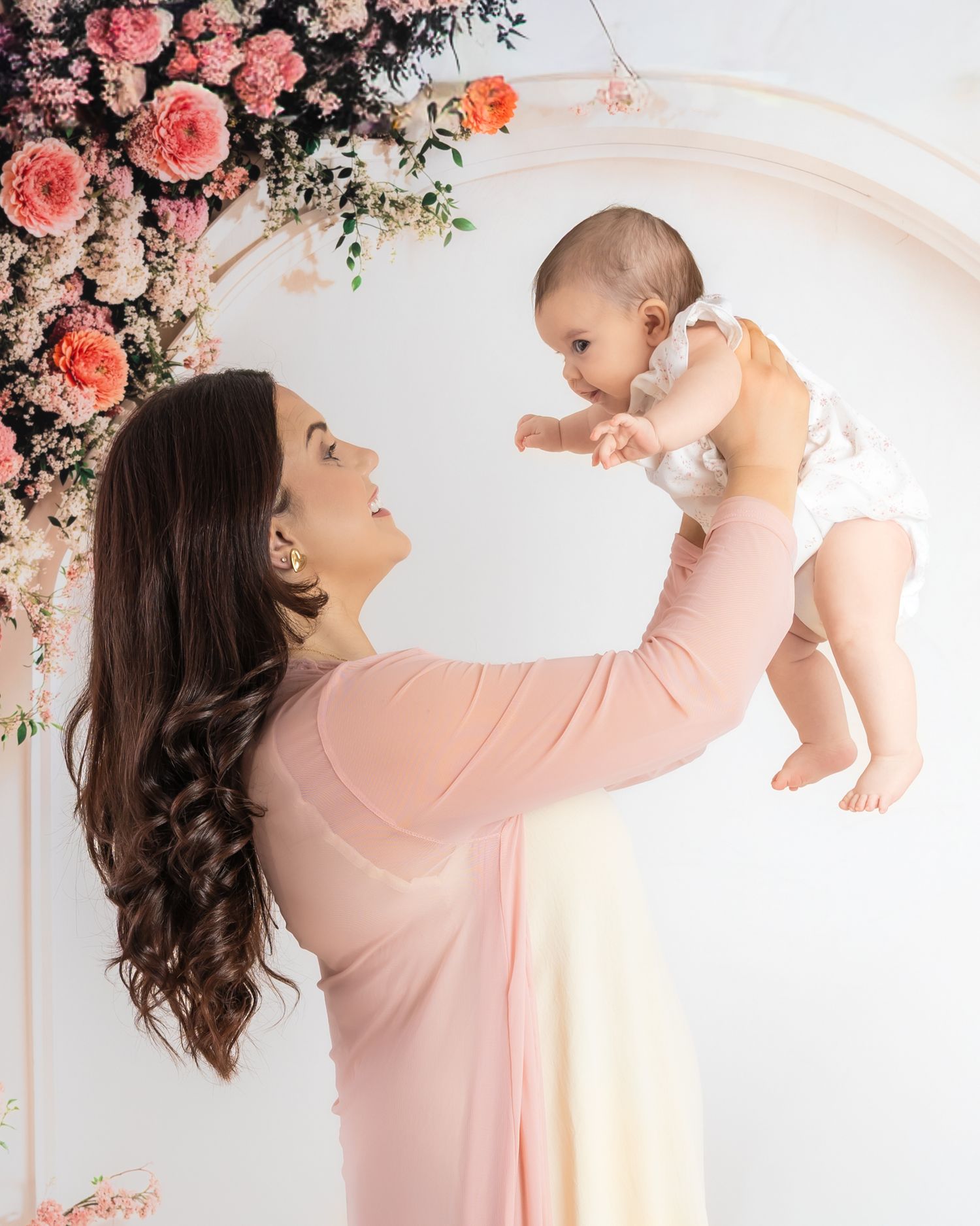 Mother in pink dress lifts baby against floral backdrop in dreamy studio portrait series.