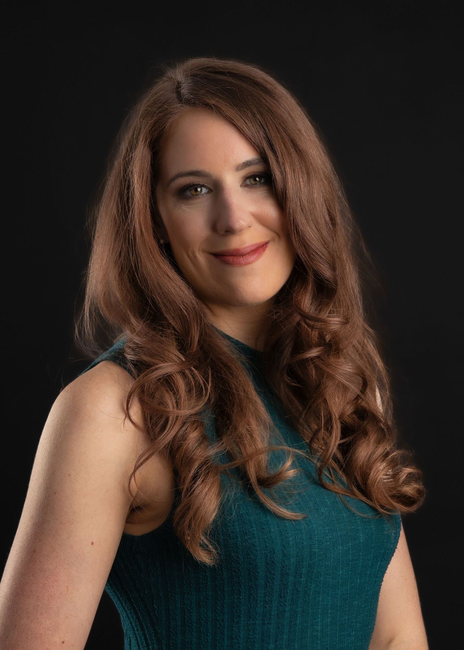 Professional headshot of a woman with long wavy auburn hair wearing a teal sleeveless top against a dark background.