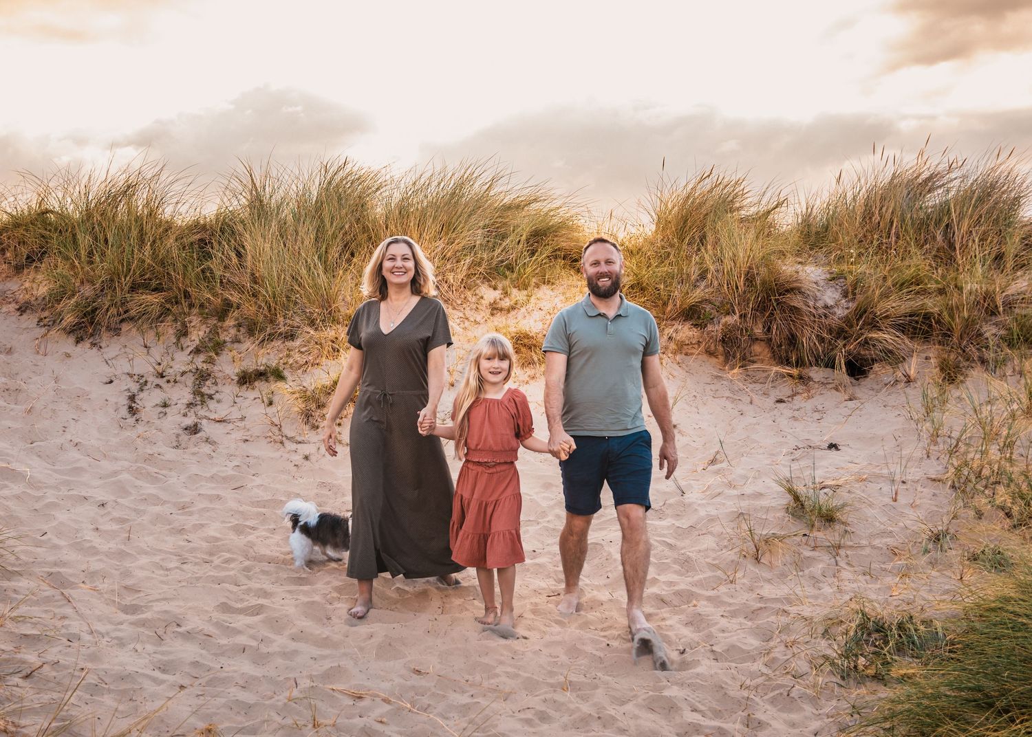 Family and small dog walking together on sandy beach dunes at sunset.