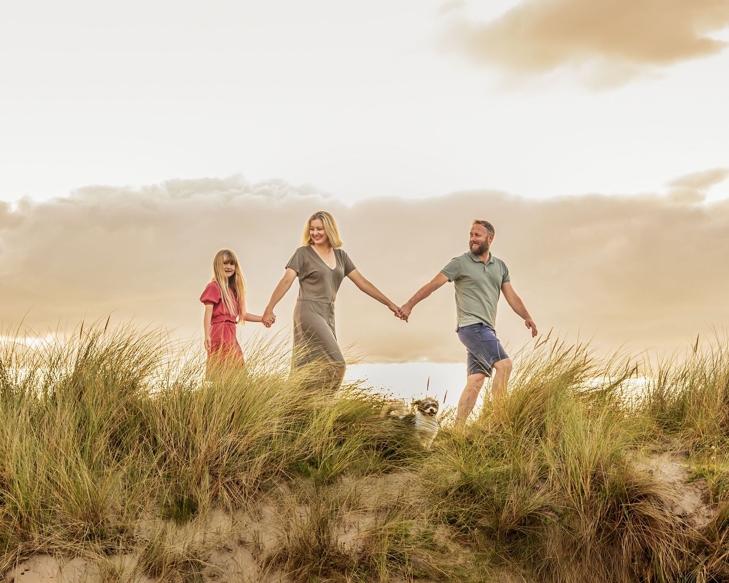 Silhouettes of people holding hands while walking along grassy sand dunes at sunset.