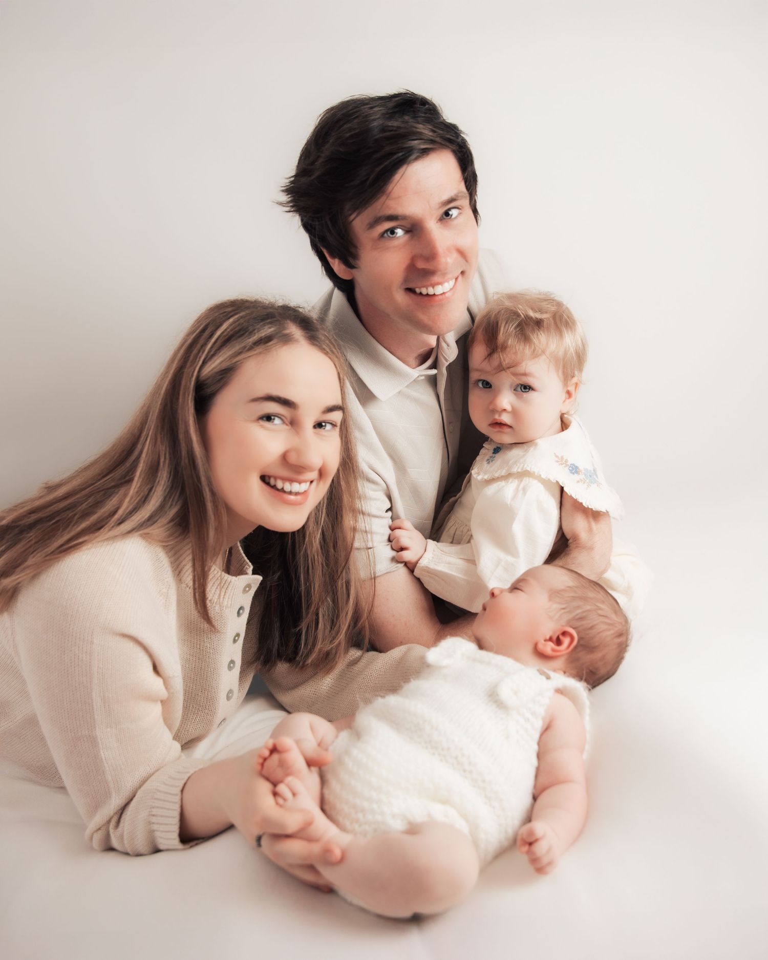 A young family poses together, parents smiling with a toddler and newborn baby, all dressed in white on a light background.