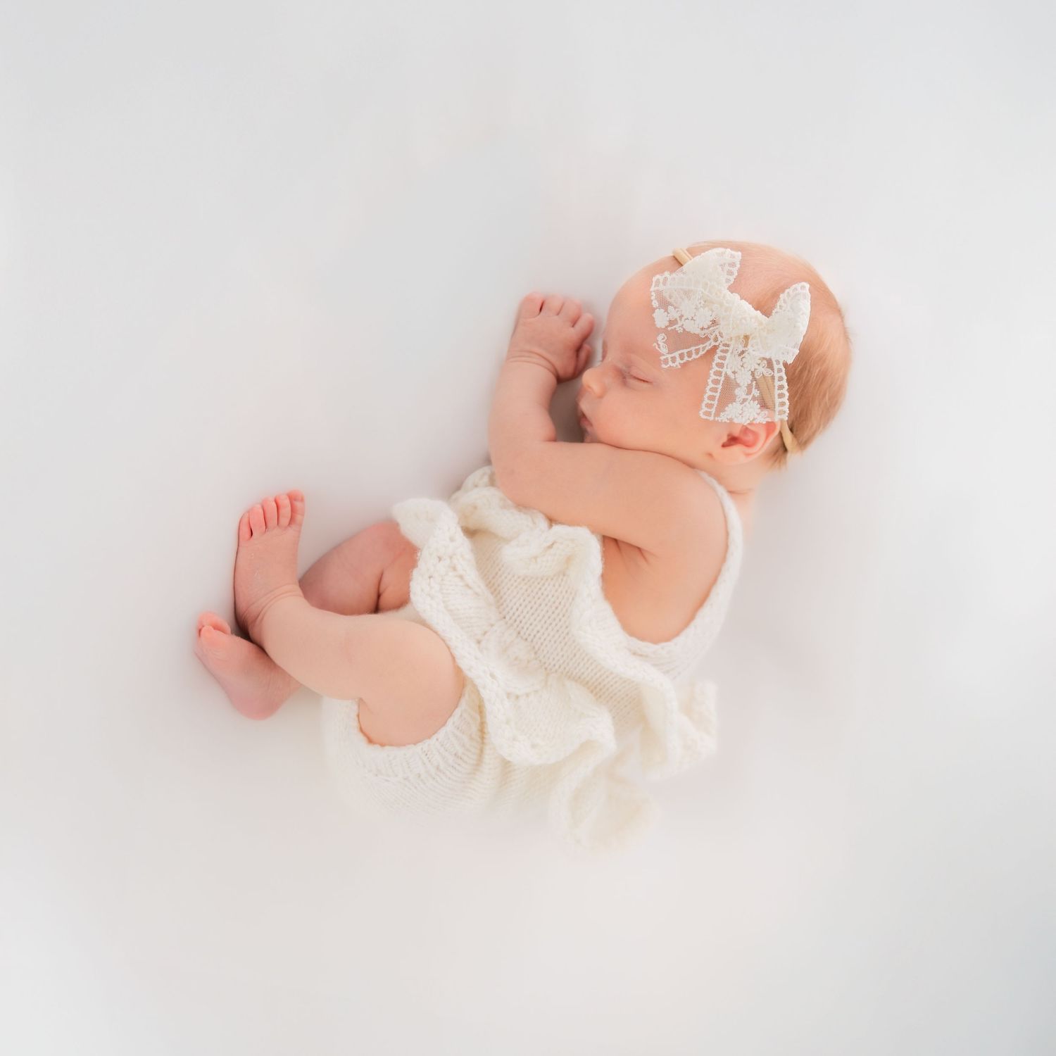 Sleeping newborn baby girl in white knit romper with bow headband on white background.