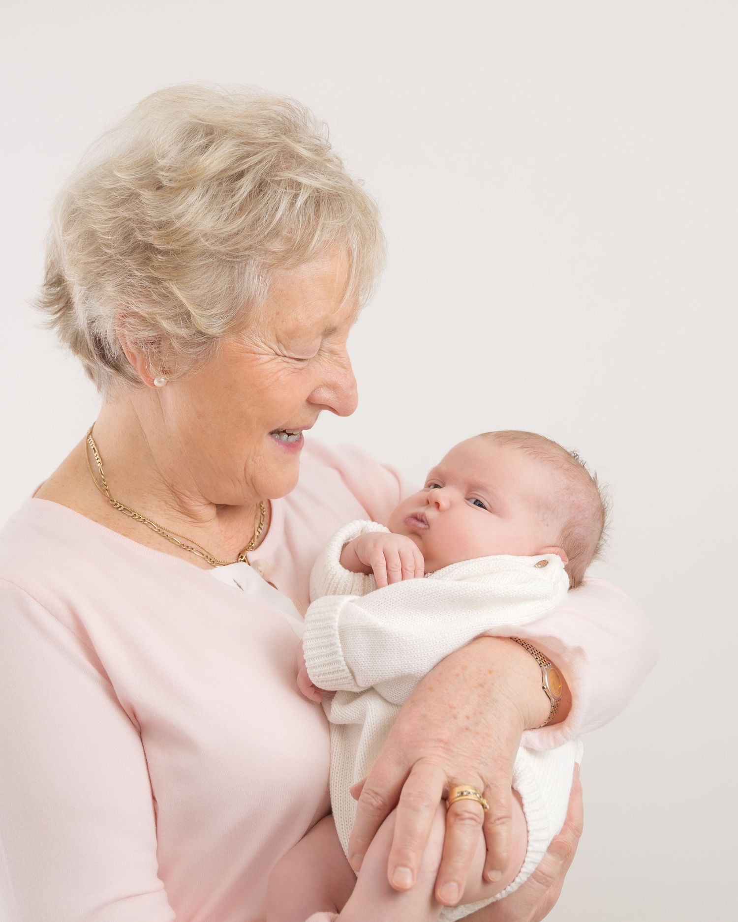 An elderly woman in pink holds and lovingly gazes at a newborn baby wrapped in white.