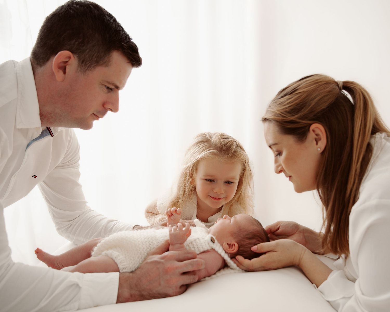 Family gathered around newborn baby on white bedding for intimate portrait.