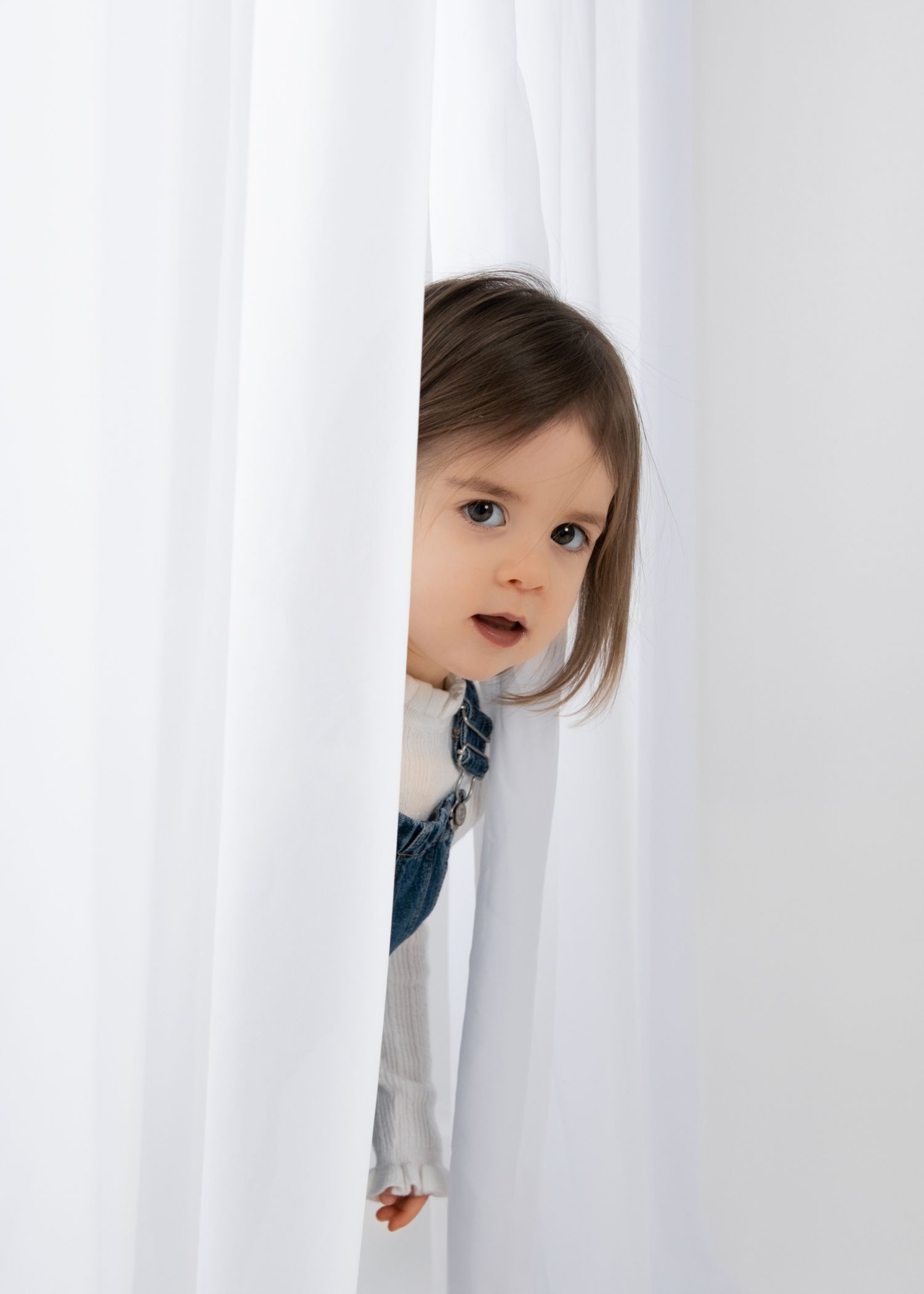 Curious toddler girl in denim overalls peeking through white curtains in a bright studio setting.