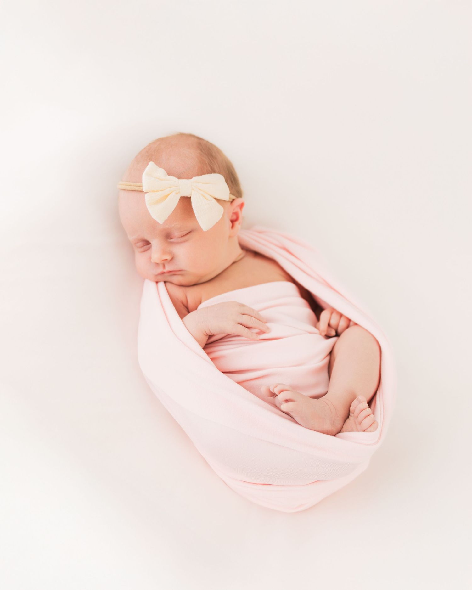 Newborn baby girl swaddled in pink wrap with cream bow headband, sleeping peacefully on white background.