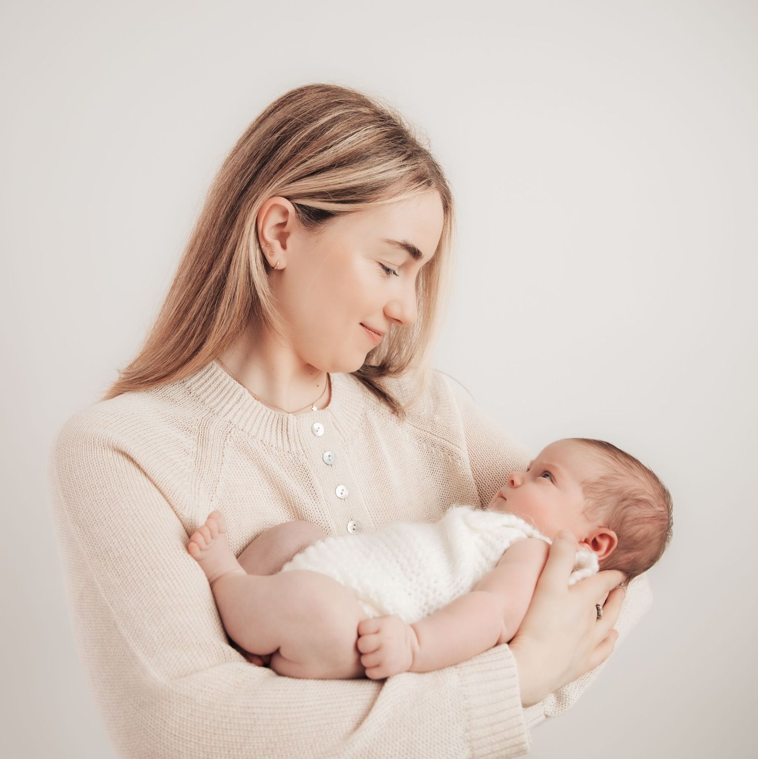 A young mother in a cream sweater tenderly holds her sleeping newborn baby wrapped in white against a soft background.