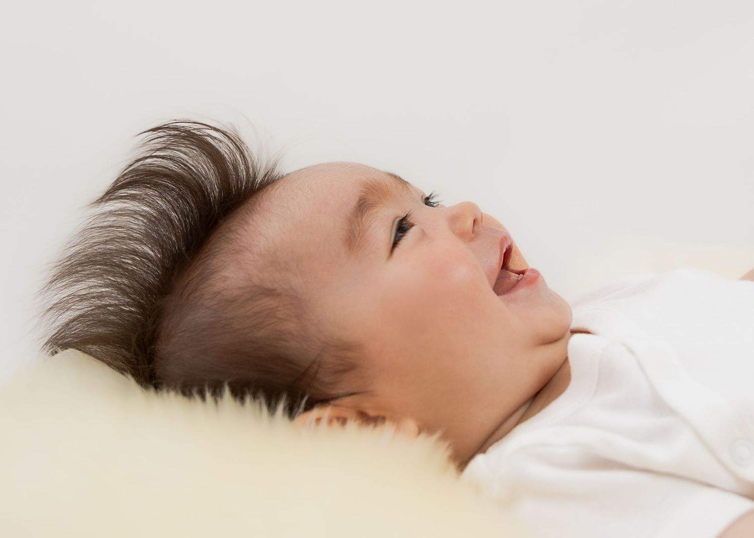 Smiling newborn baby with dark hair lying on a soft yellow pillow in a white onesie.