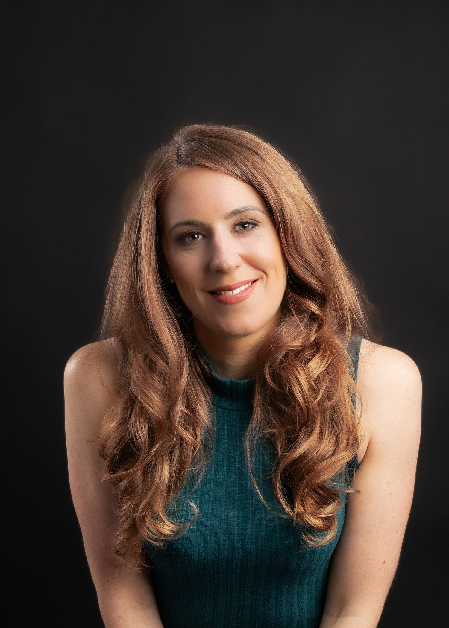 Smiling woman with long auburn hair wearing a teal sleeveless top against a dark studio background.