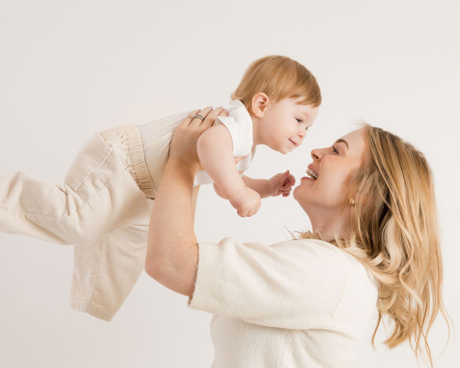 A parent in cream-colored clothing lifts and plays with a happy baby against a light meatyh studio background.