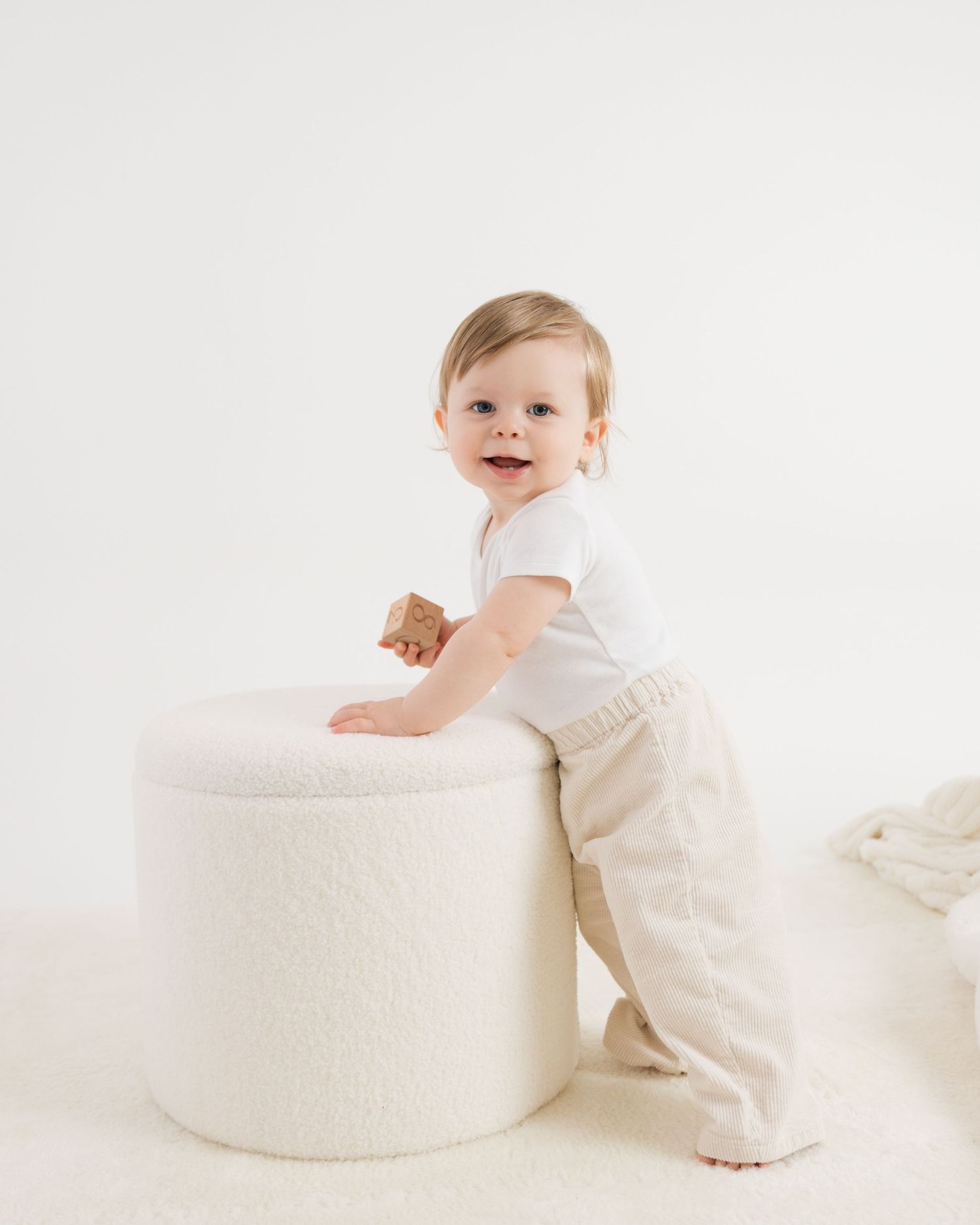 A baby in white clothing crawls and plays around a round cream-colored ottoman or pouf in a bright home meath studio setting.