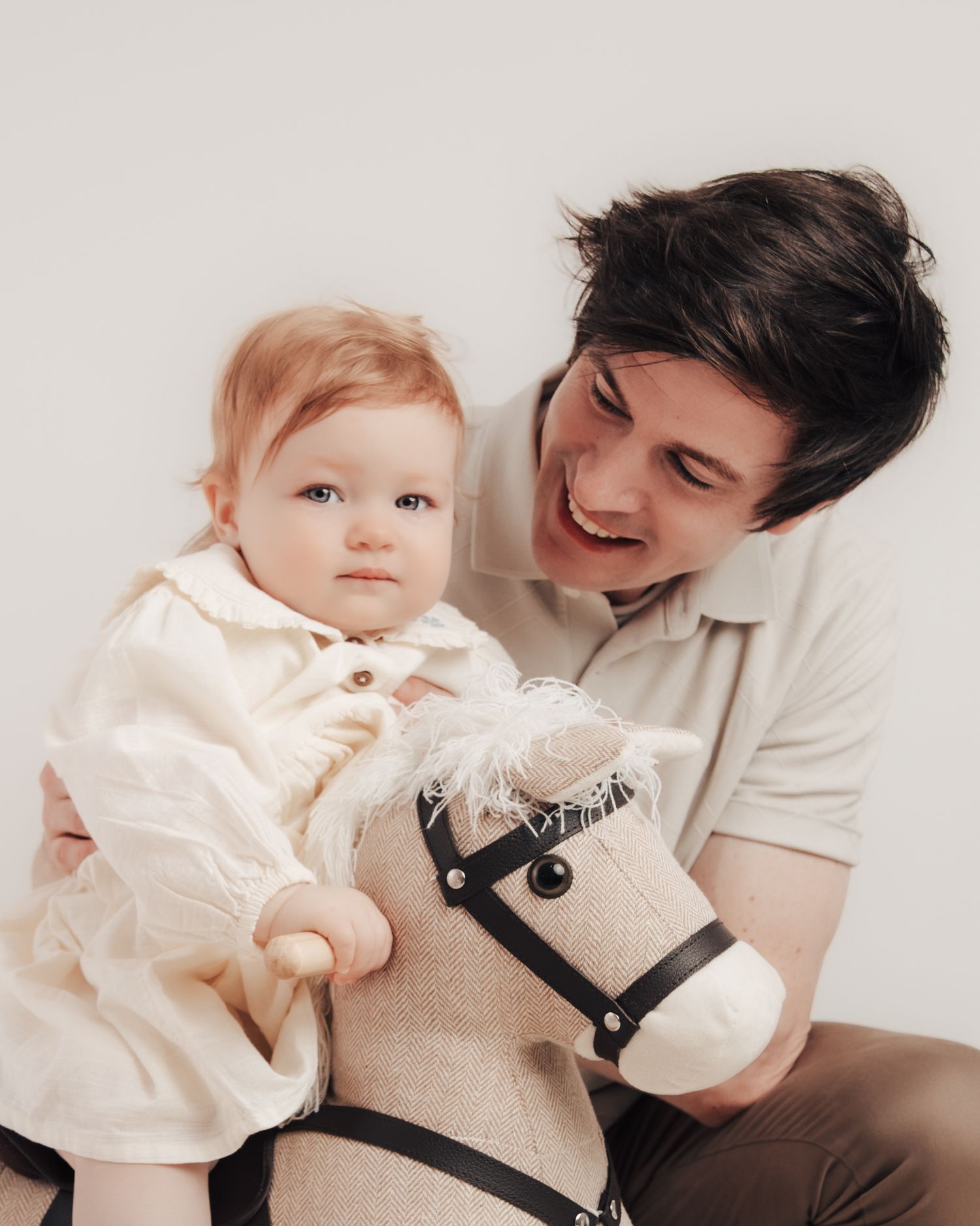 A smiling man holds a red-haired baby in a white dress, both posing with a toy hobby horse.