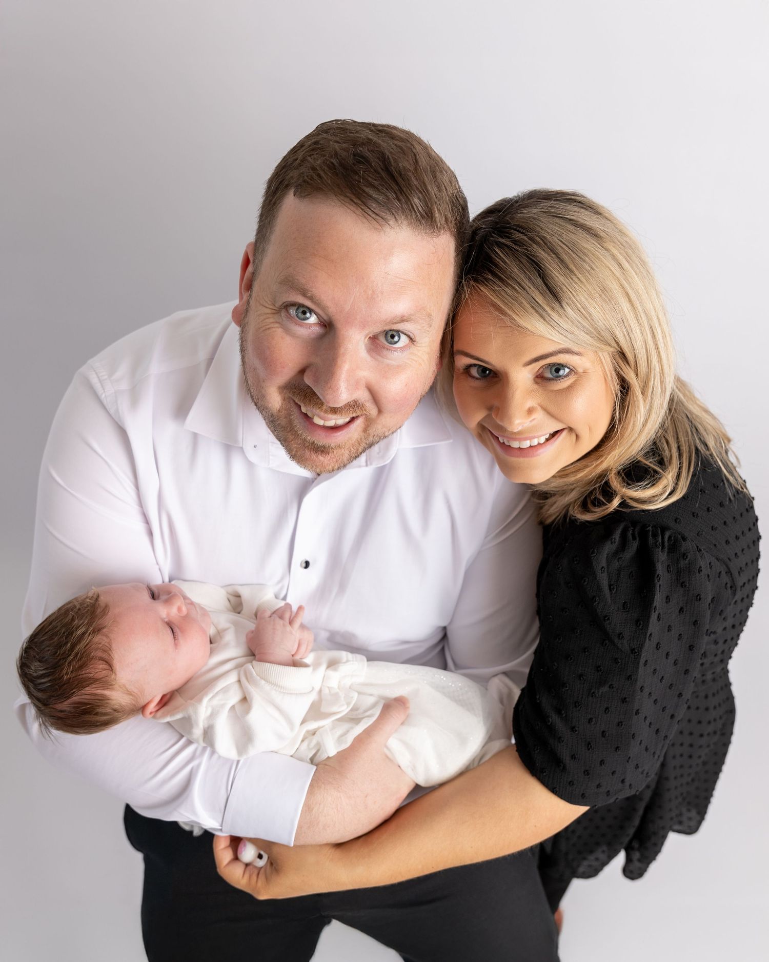 A family portrait with parents holding their newborn baby against a gray background.