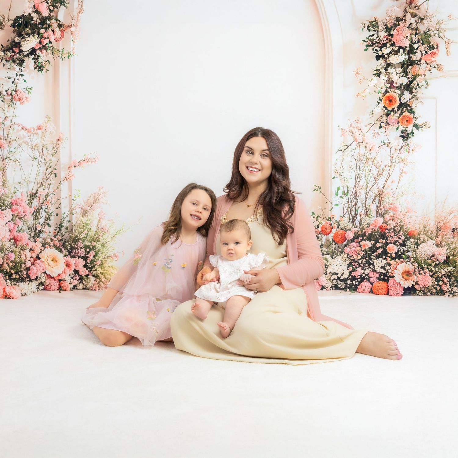 Mother and daughters in pink dresses sit together on white floor surrounded by delicate floral arrangements.