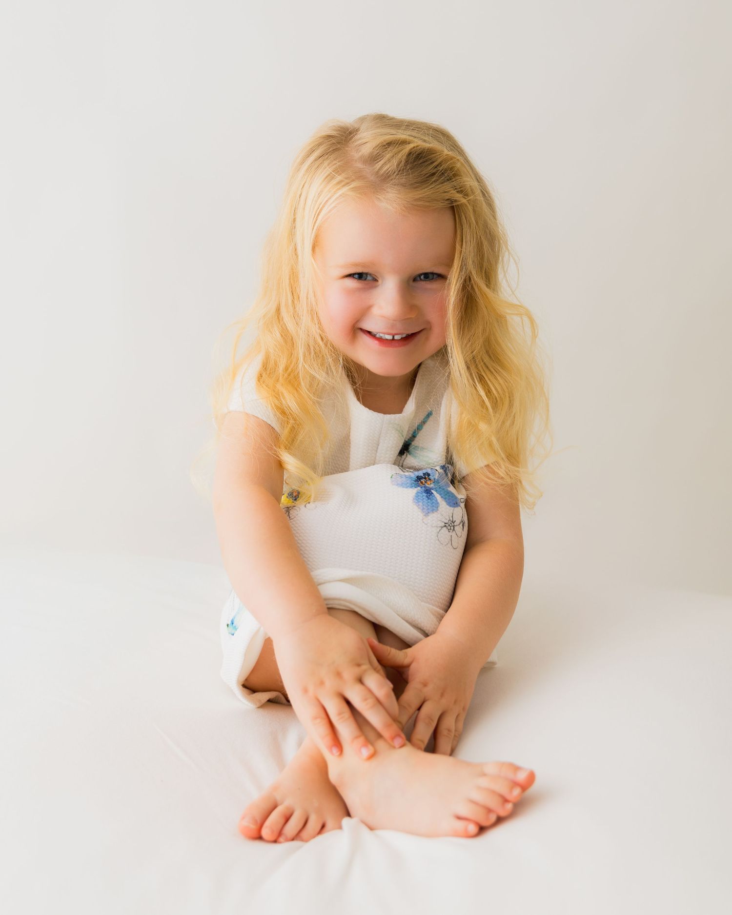 Smiling young blonde girl in white floral outfit sitting barefoot on white background in a studio portrait.