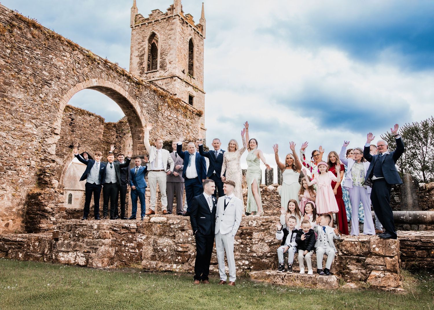 Large wedding party celebrating among ancient stone church ruins with raised arms under a cloudy sky.