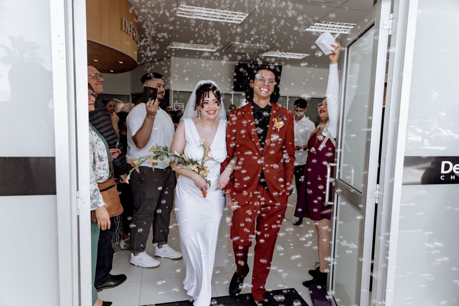 Group of wedding guests throw confetti in celebration inside an indoor venue hallway as newlyweds walk through.
