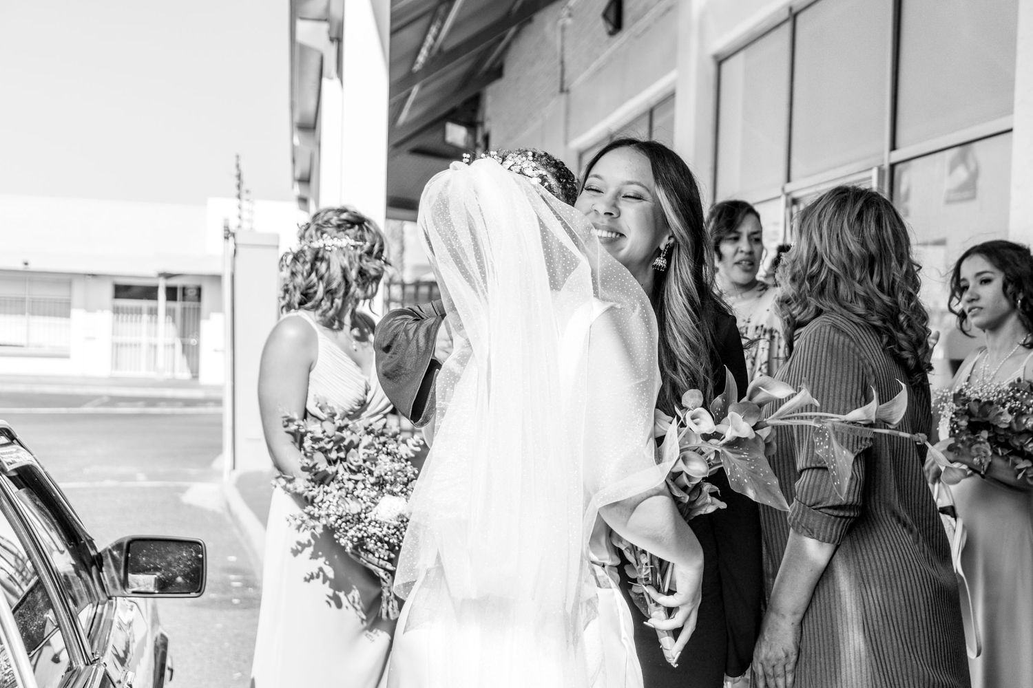 A joyful wedding party gathered outside on a sunny day, with elegant floral bouquets and formal attire.