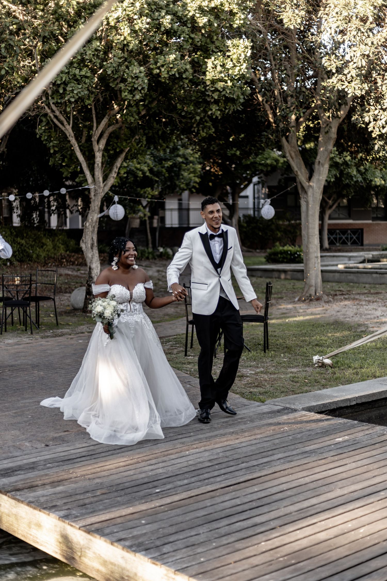 Elegant wedding couple walking together on a wooden deck surrounded by trees in a garden setting.