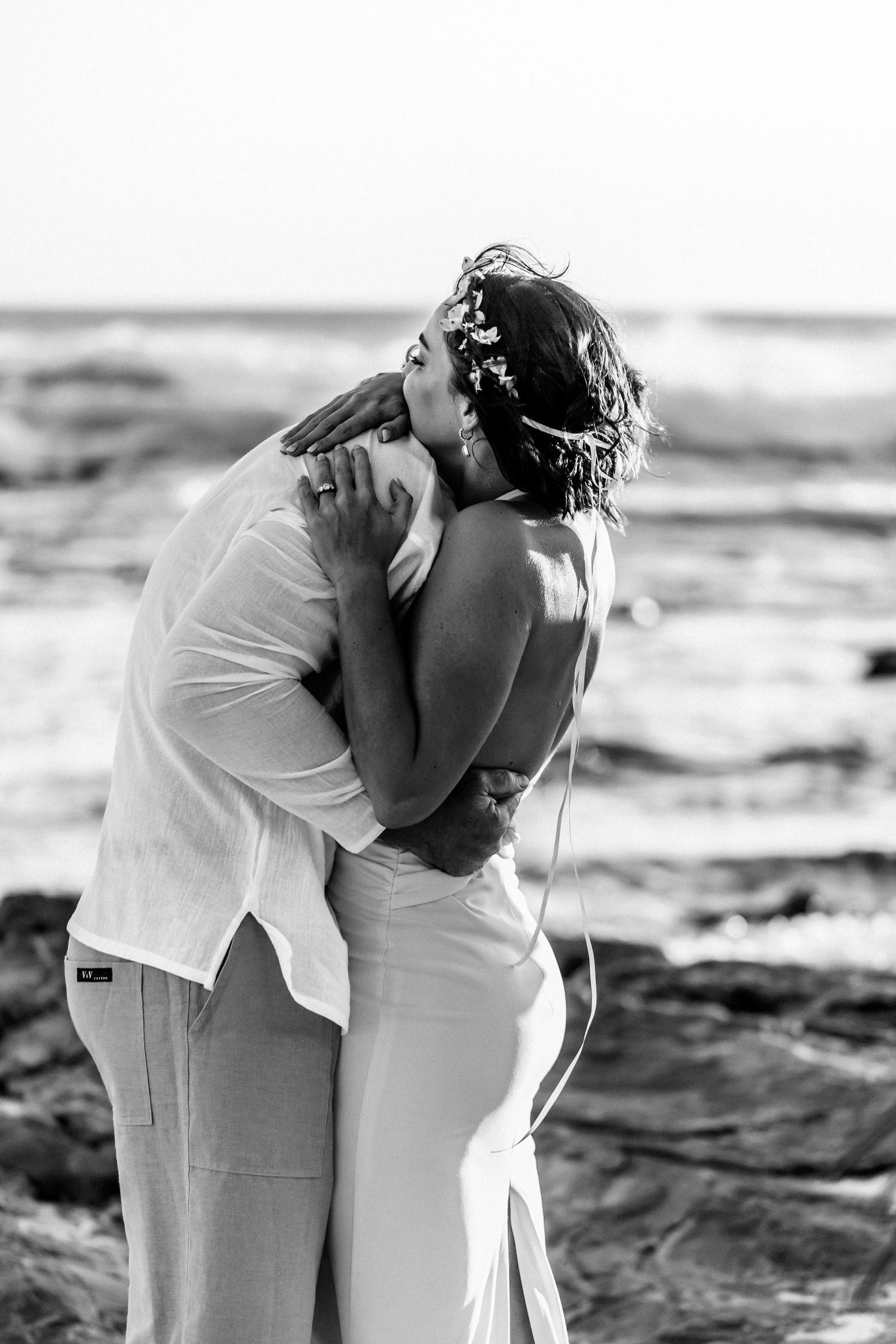 A romantic black and white beach wedding photo shows an intimate embrace on rocky coastal shores.