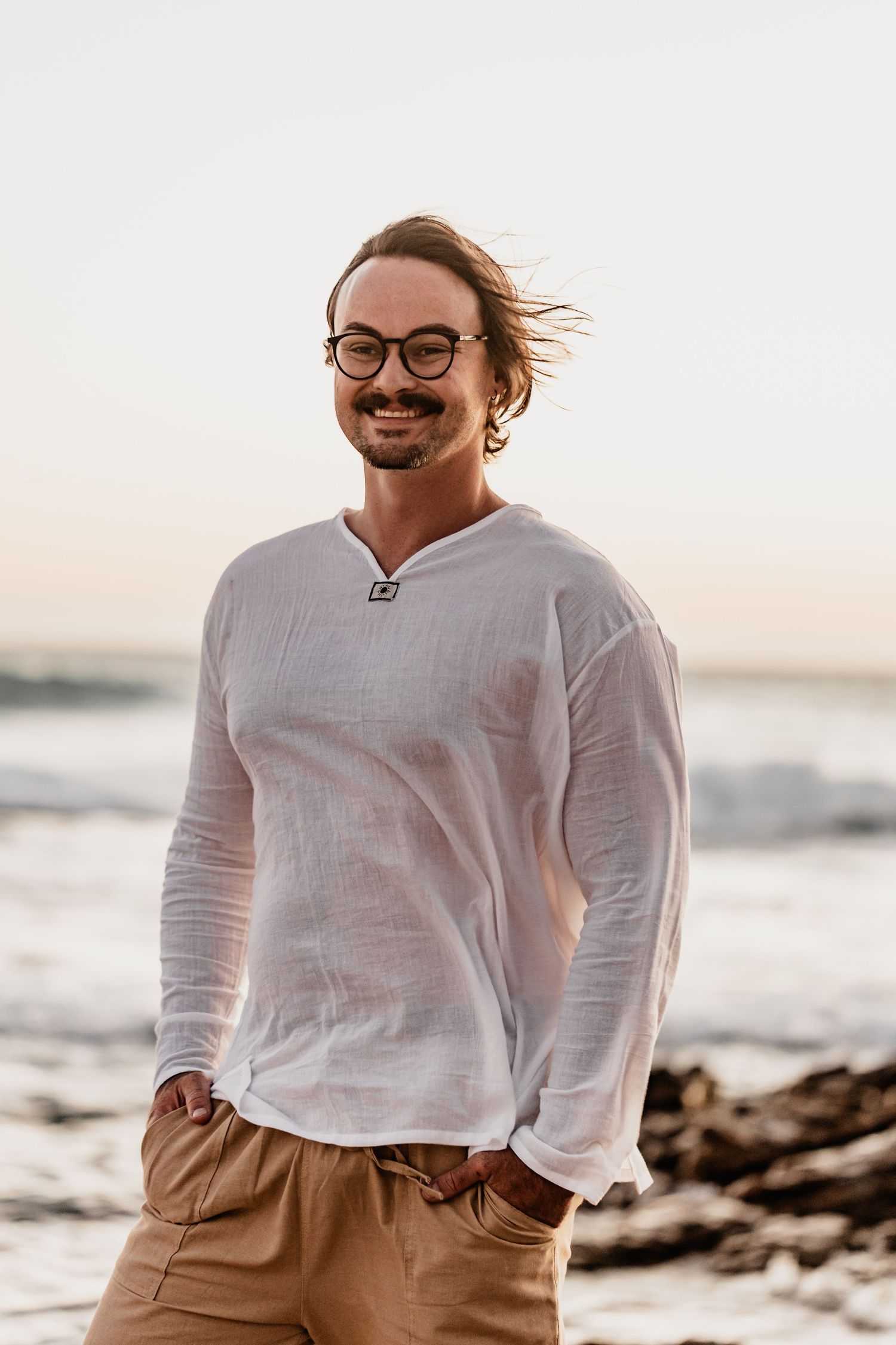 Breezy beach portrait of a smiling person in glasses wearing a white long-sleeve top against a coastal sunset backdrop.