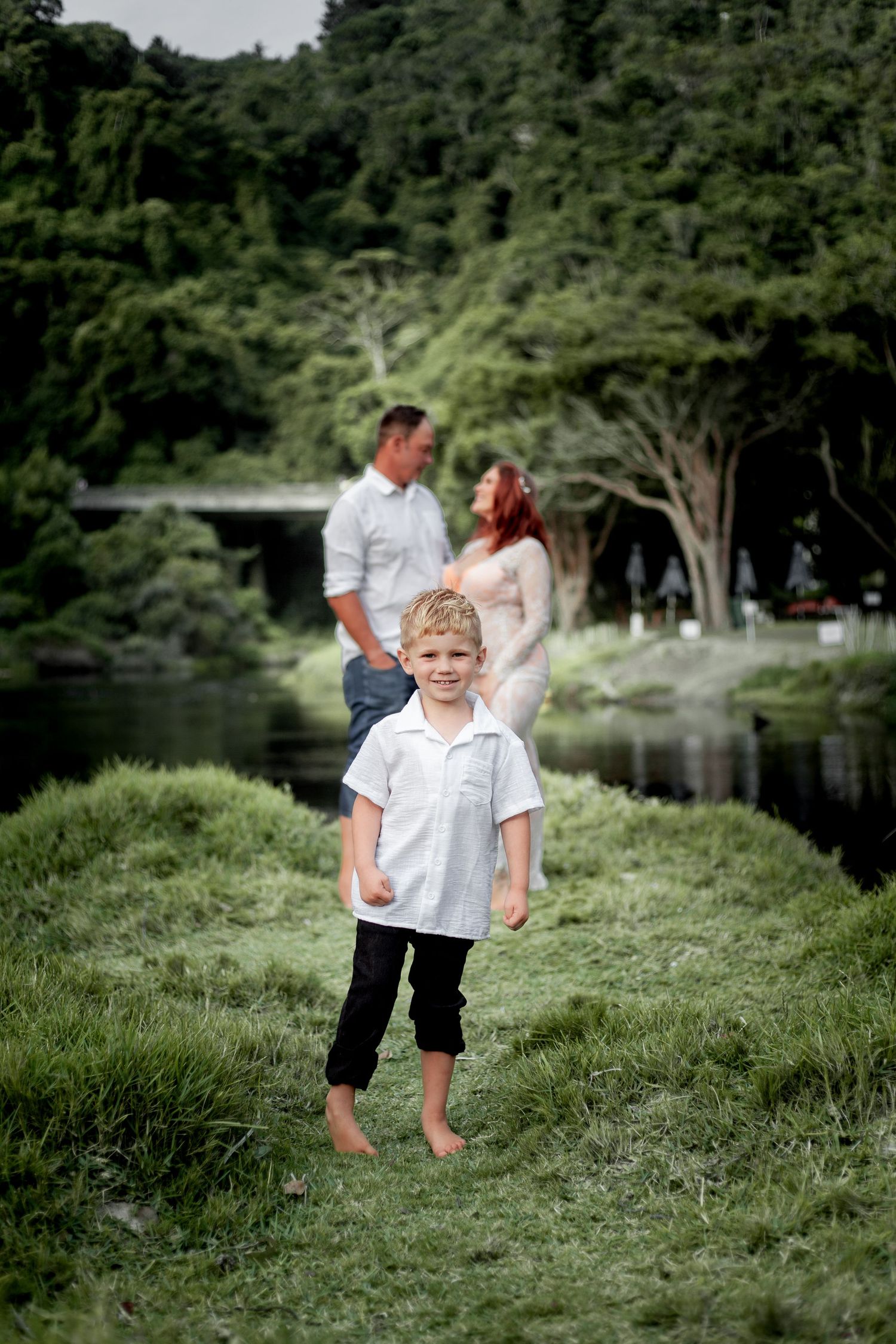 A young child stands barefoot on grassy terrain near a tranquil pond with tall trees in the background.