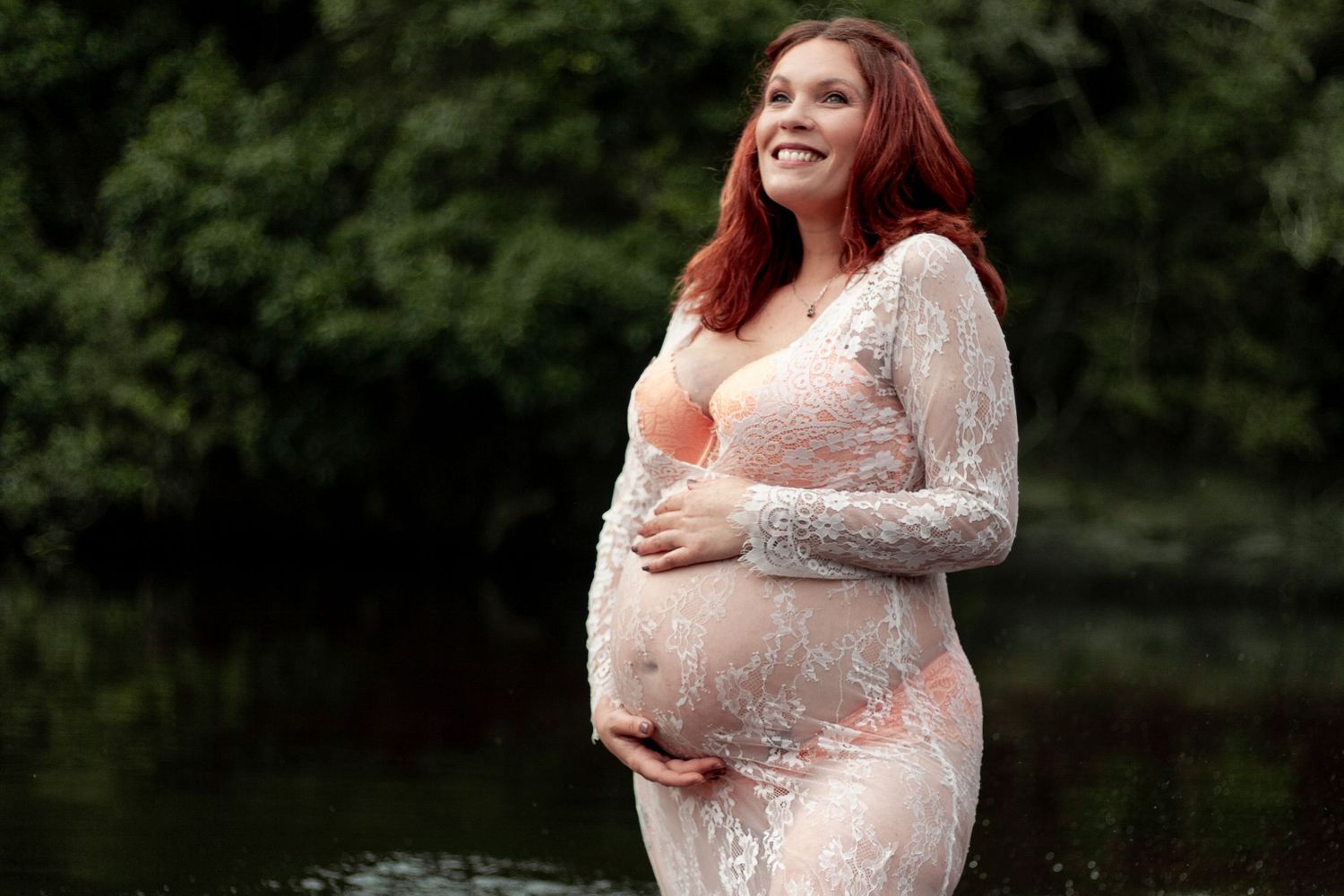 Expectant mother wearing white lace dress poses near calm water during maternity photo session outdoors.