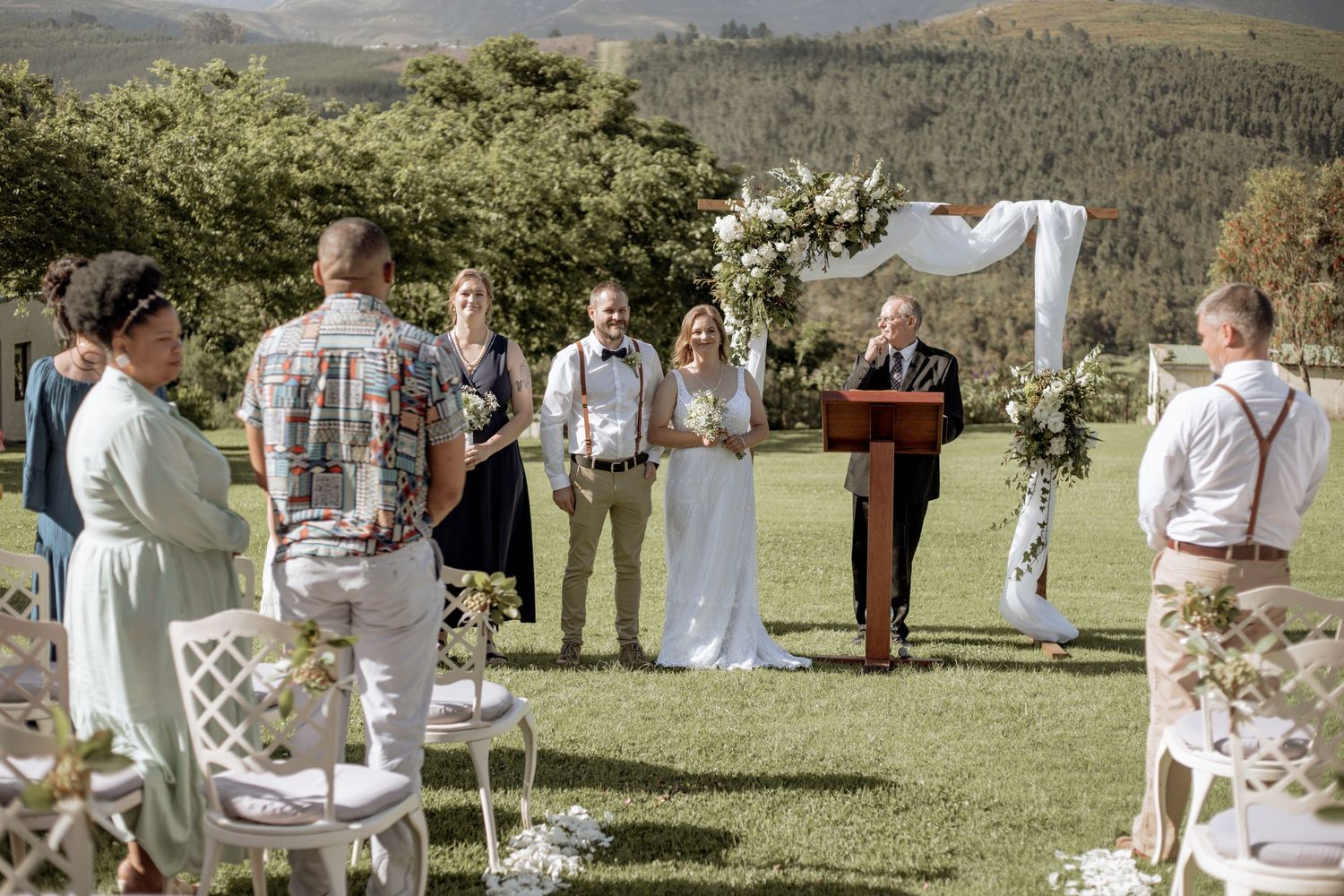A panoramic view of an elegant outdoor wedding ceremony with white-clad guests gathered in a scenic mountain setting.