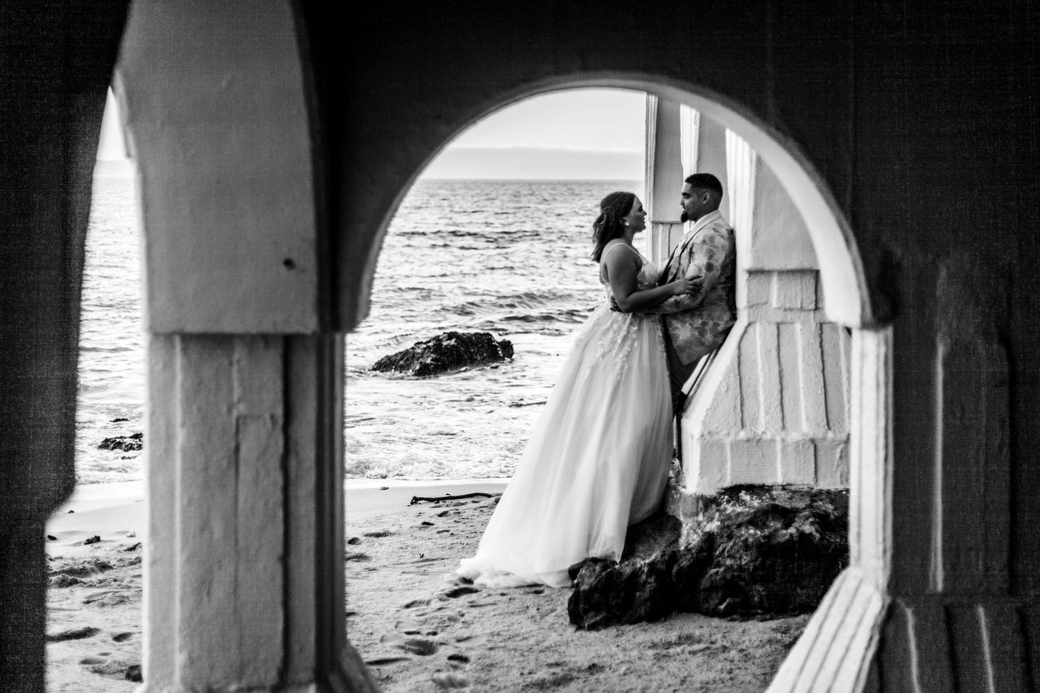 A couple in wedding attire embraces beneath an archway overlooking a rocky beach in black and white.