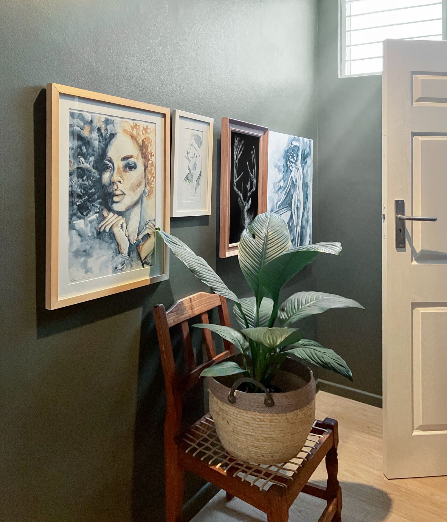 A modern hallway gallery wall displays framed artwork alongside a wooden chair and potted plant against sage green walls.