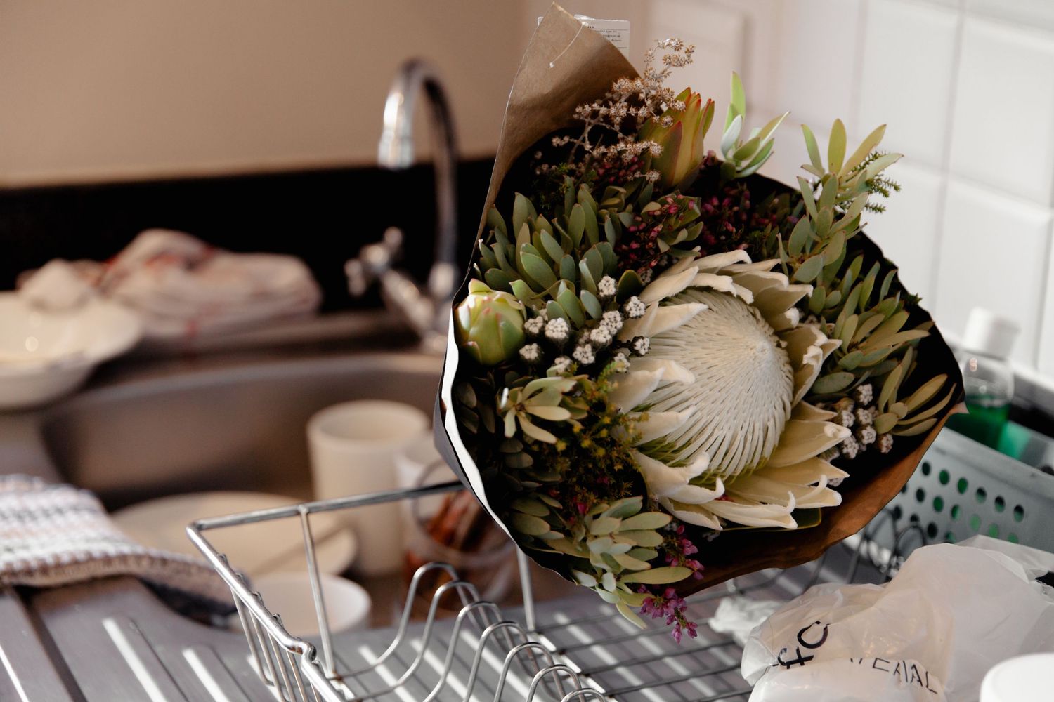 A native floral bouquet with protea and greenery rests on a kitchen sink next to dish drying rack.