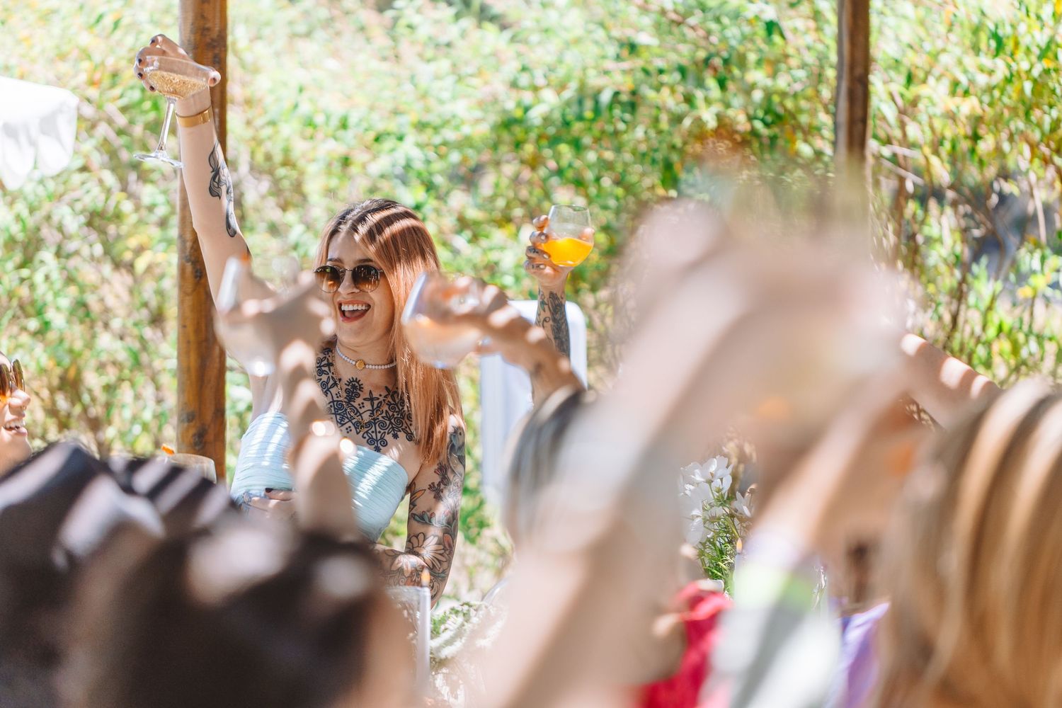 Woman raising glass at outdoor festival celebration with crowd cheering in sunny garden setting.