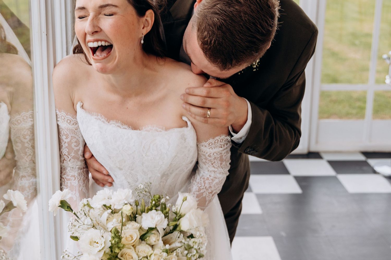 Joyful bride laughing as groom kisses her shoulder, holding white floral bouquet in elegant wedding attire.