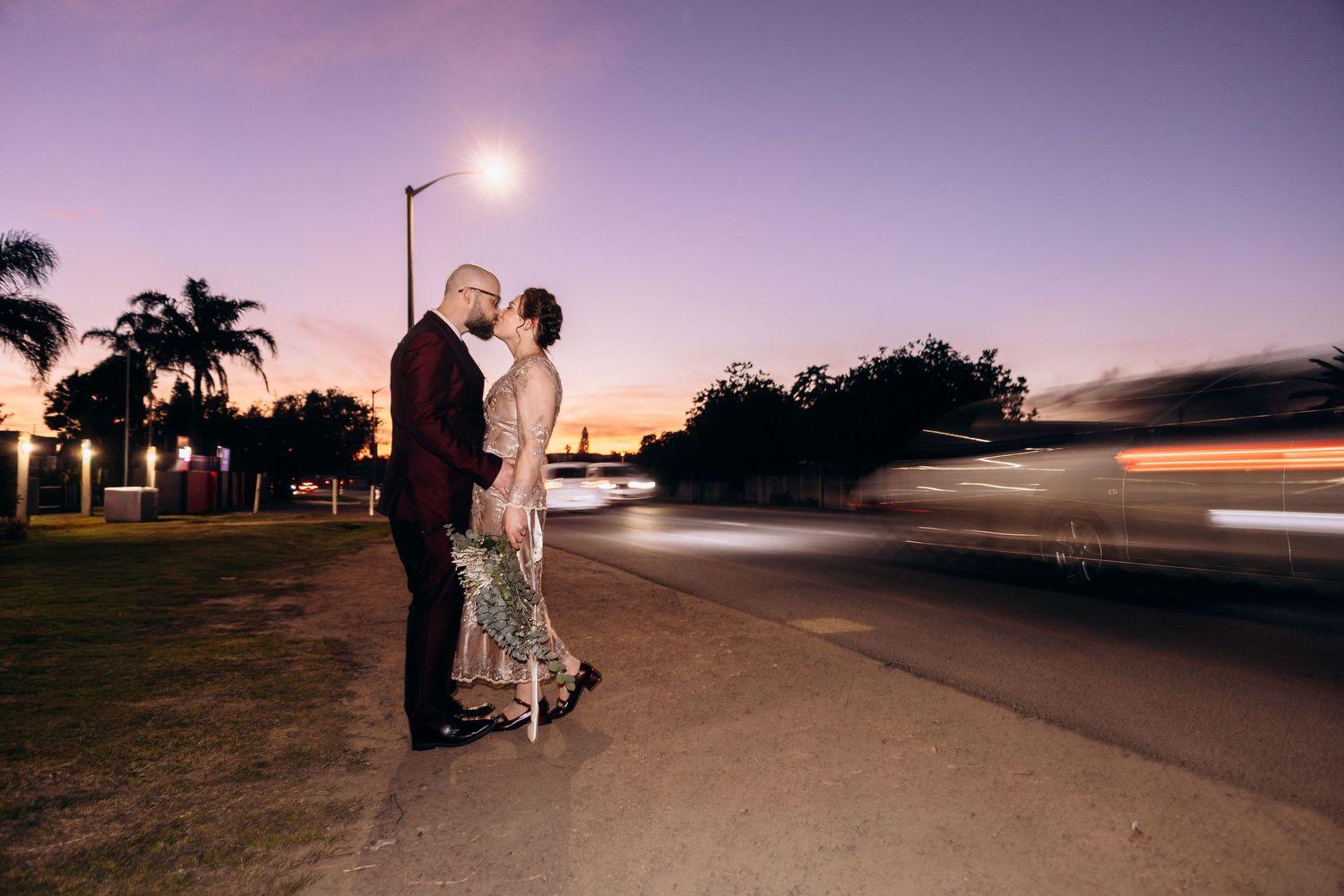 Couple embracing at dusk near a road with light trails, palm trees, and a purple sky in the background.