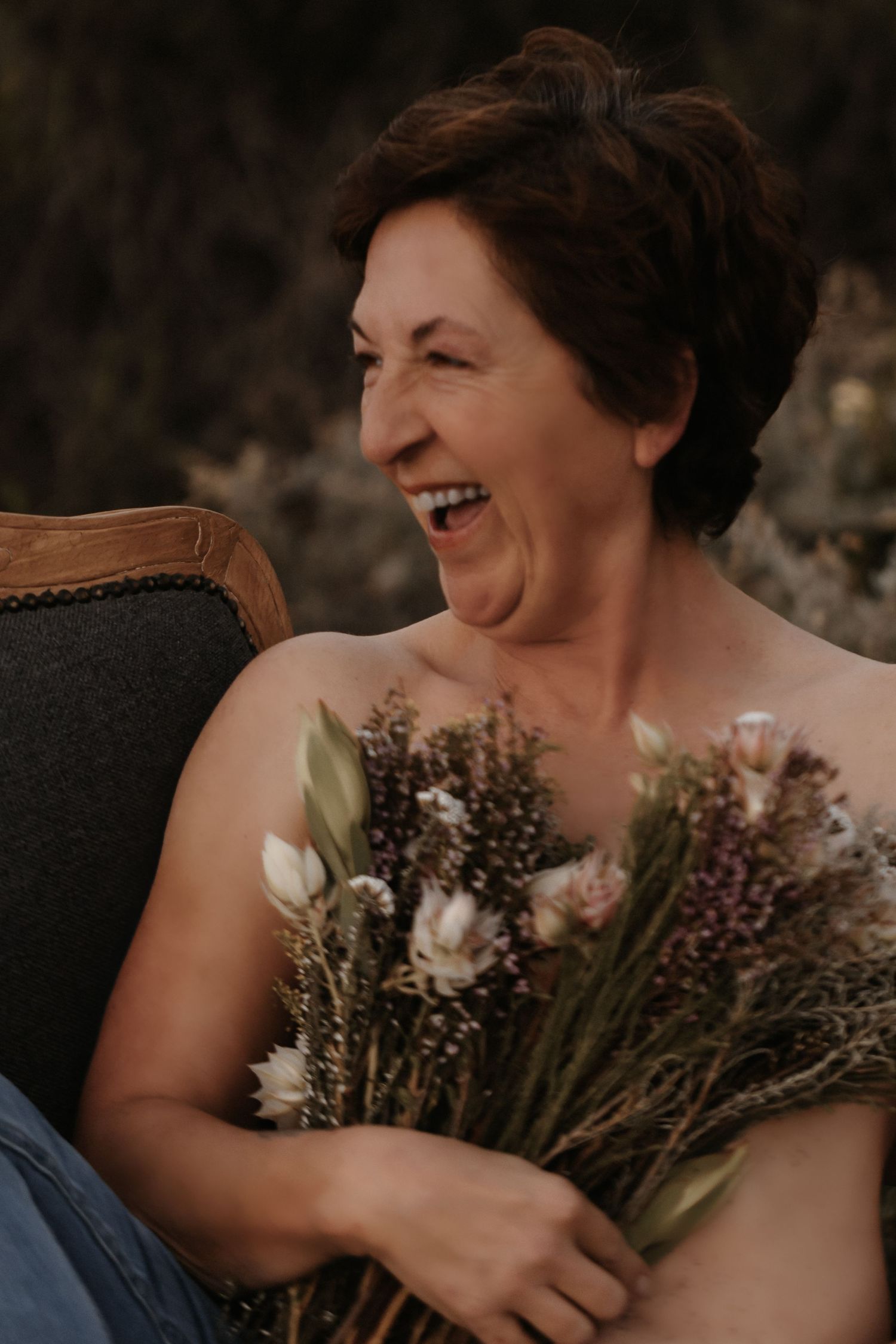 A joyful candid portrait shows genuine laughter while holding a rustic bouquet of dried flowers and tulips on an antique chair.