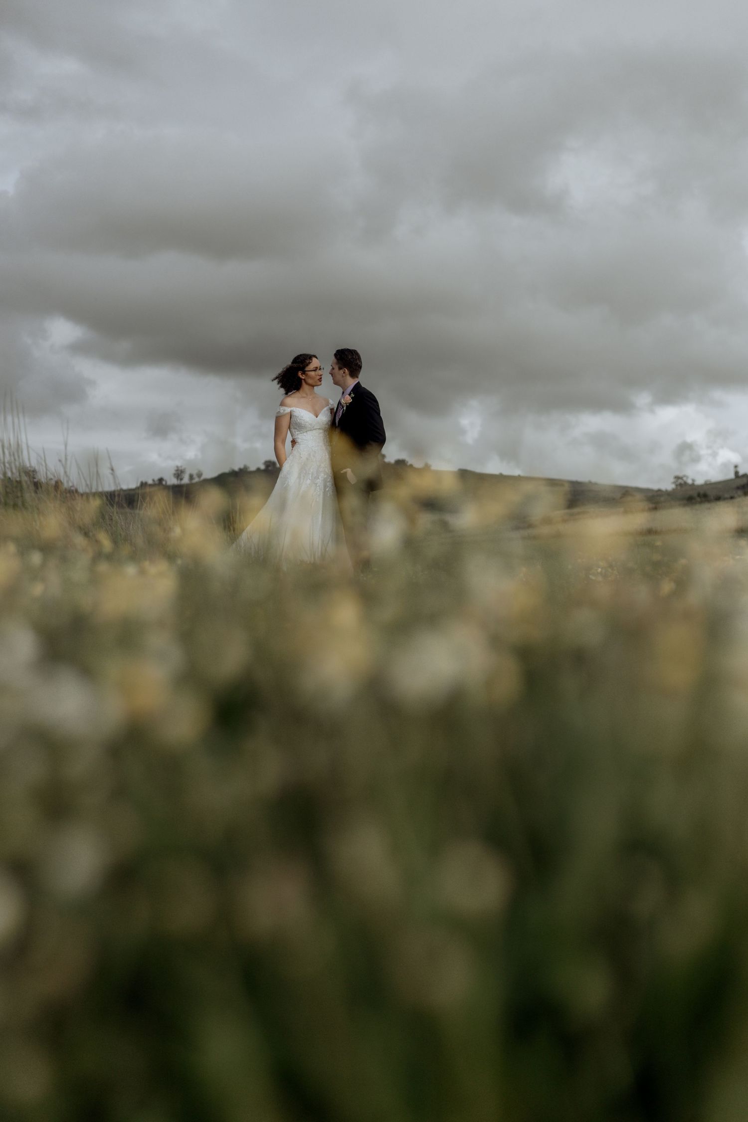 A couple stands together in a dreamy field with moody storm clouds overhead in a dramatic wedding photograph.