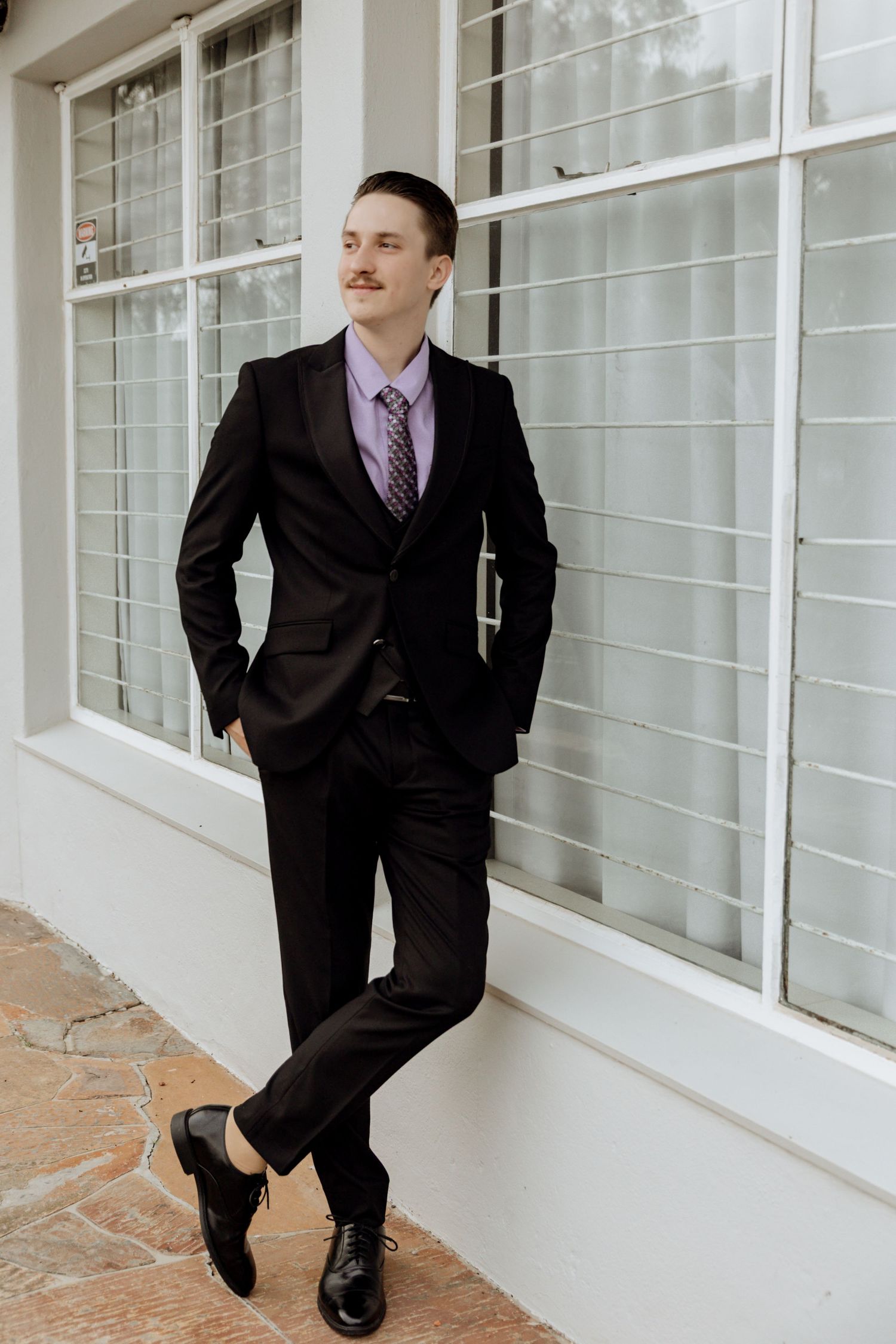 Professional wearing a dark suit and tie poses against a white exterior wall in various relaxed stances.