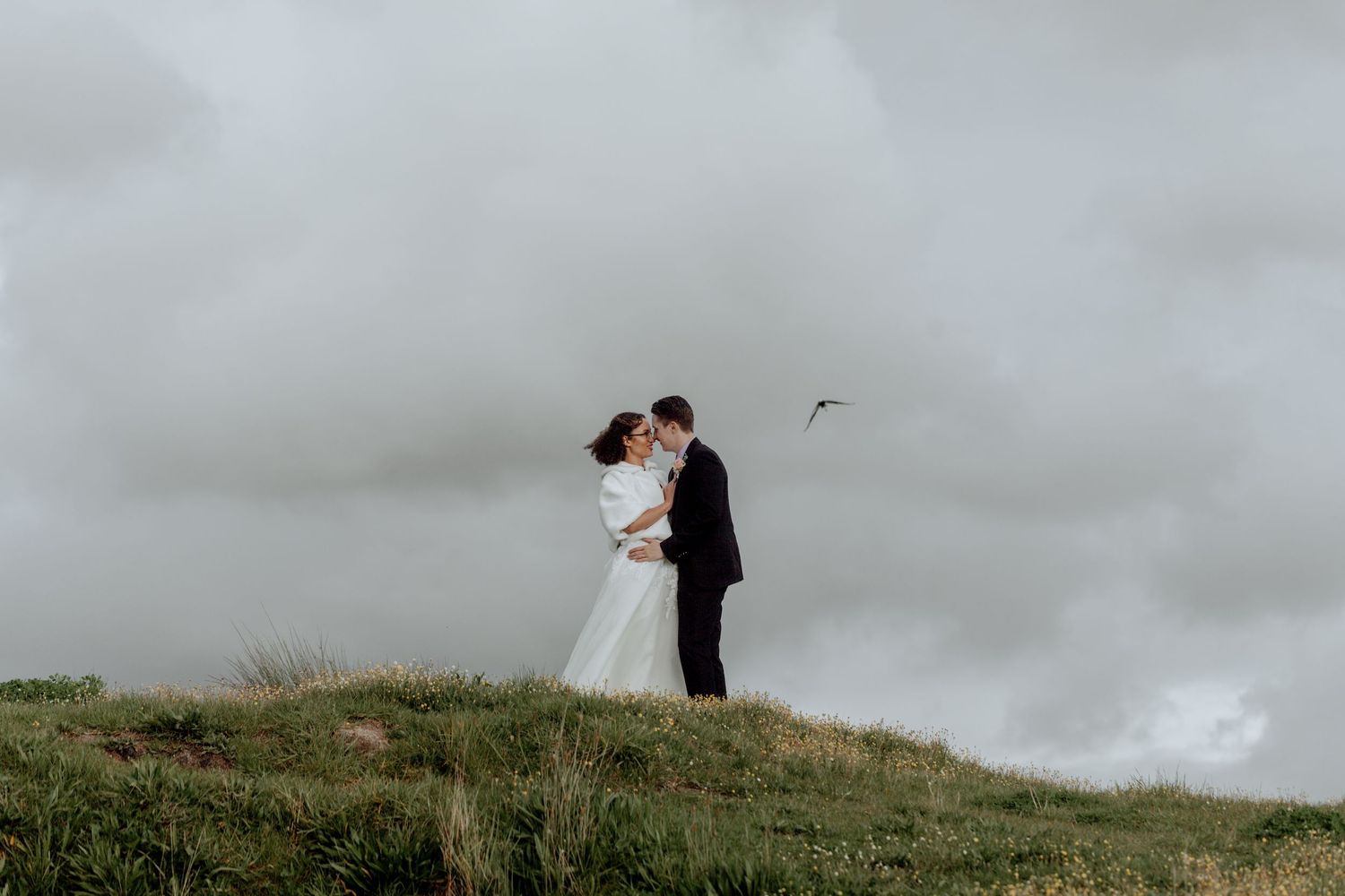 Couple stands together on a grassy hilltop silhouetted against moody clouds at sunset on their wedding day.