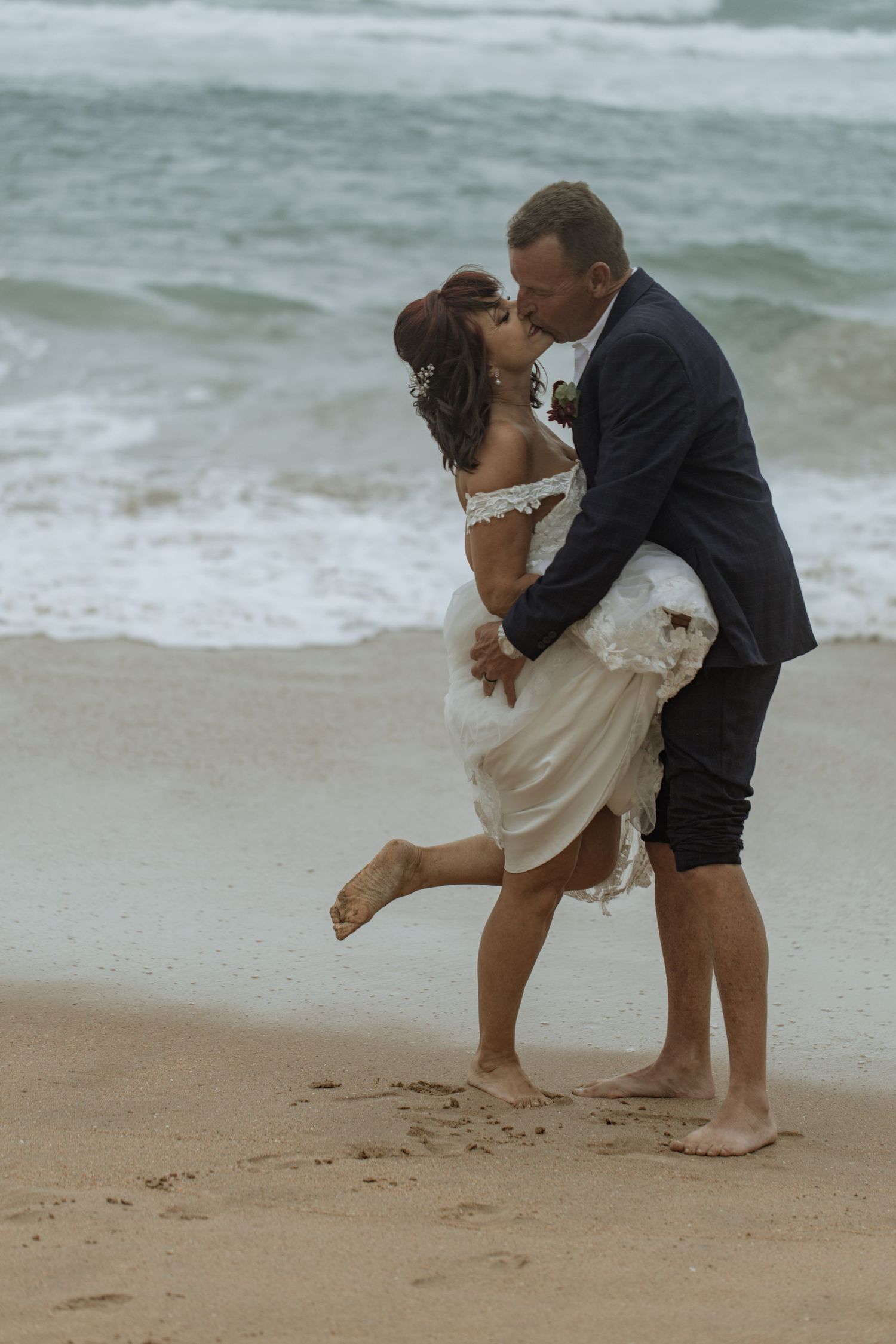A couple in formal attire shares a romantic moment while drawing in the sand on a cloudy beach.