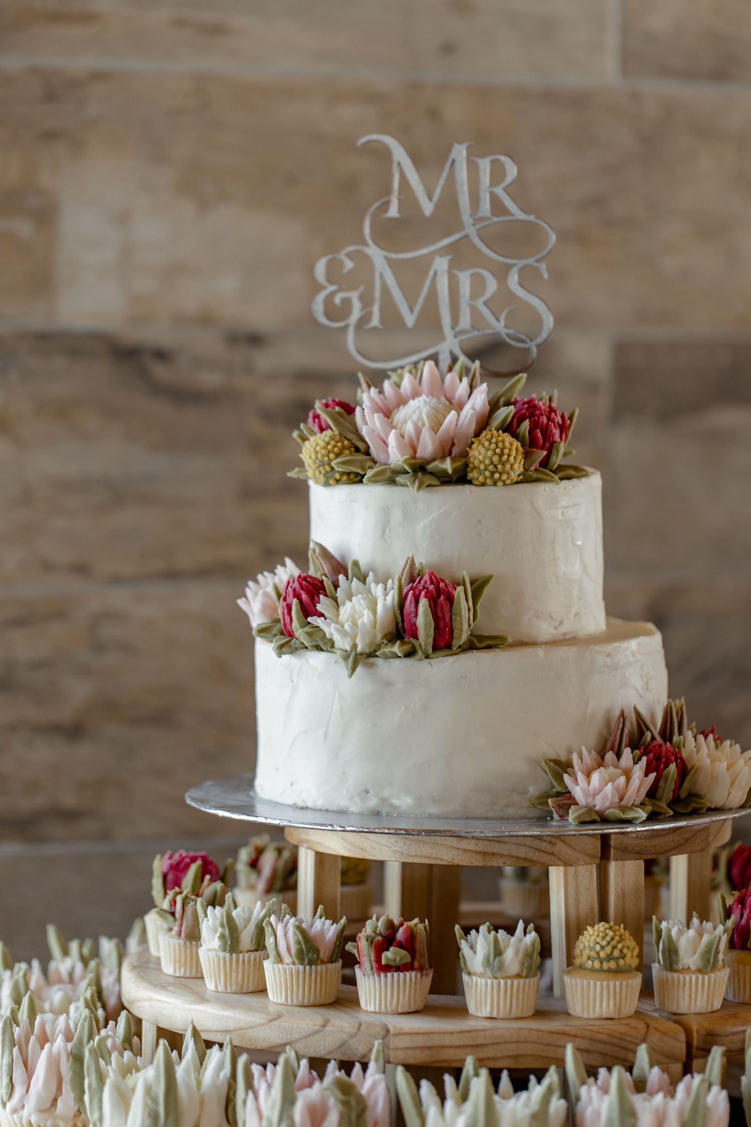 White wedding cake with fresh flowers and cupcakes displayed on rustic wooden tiered stand at reception.
